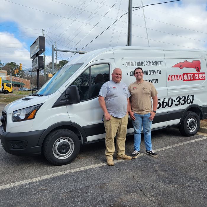 Two men are standing in front of a white van that says auto glass