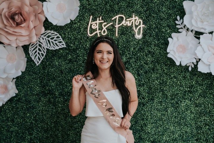 Woman in white dress and sash smiling at a bachelorette party backdrop with flowers and “Let’s Party” sign