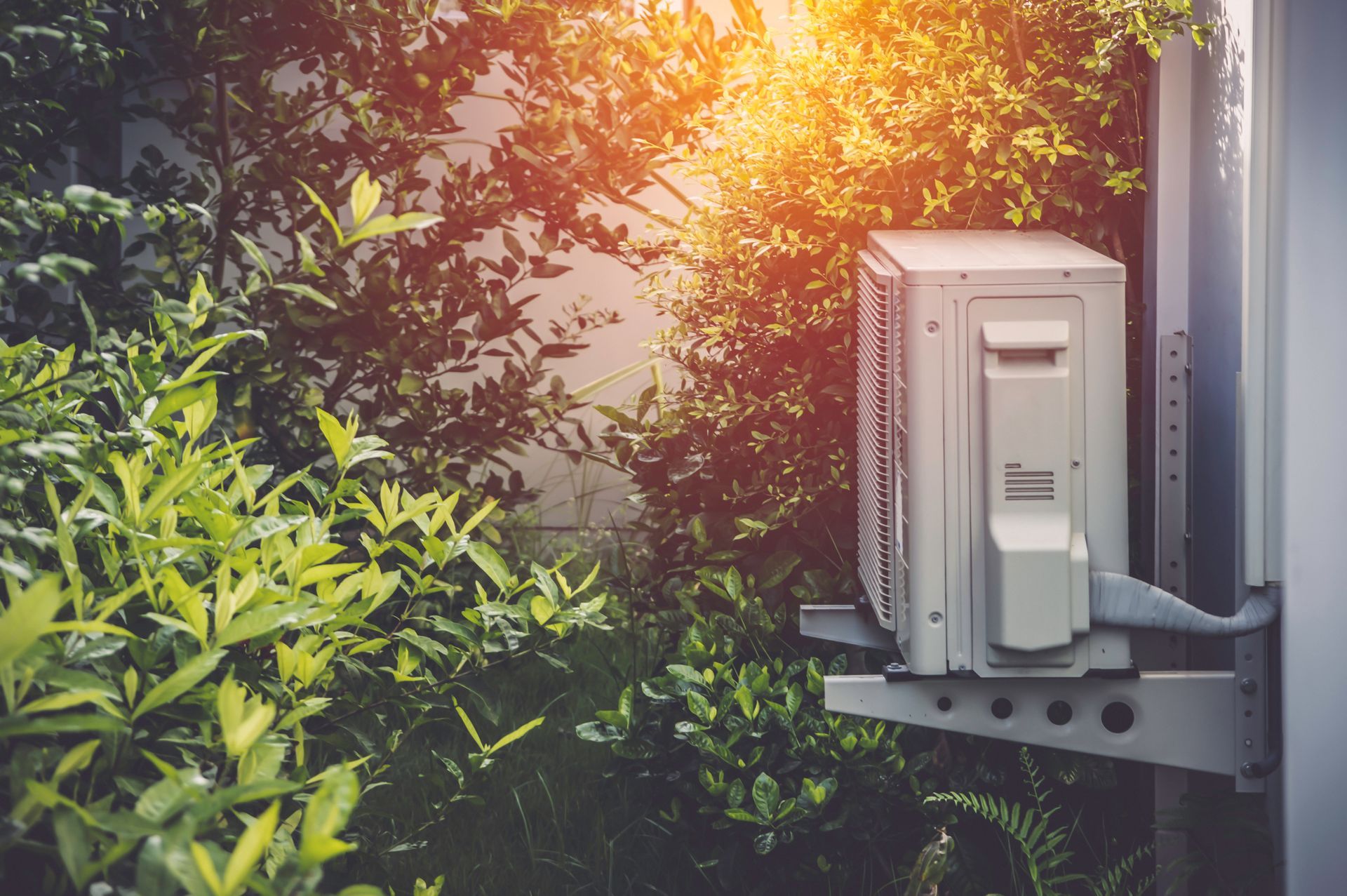 White air conditioning unit mounted outside, surrounded by green foliage, sunlit background.