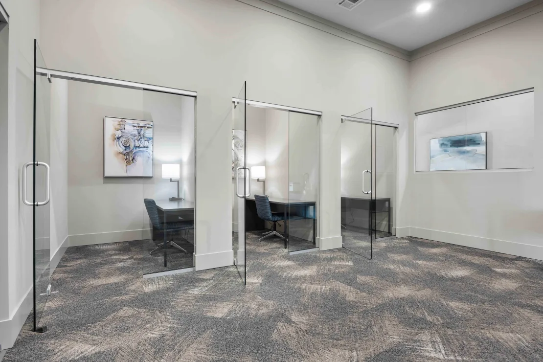 Glass-walled private offices with desks and chairs in a modern community workspace.