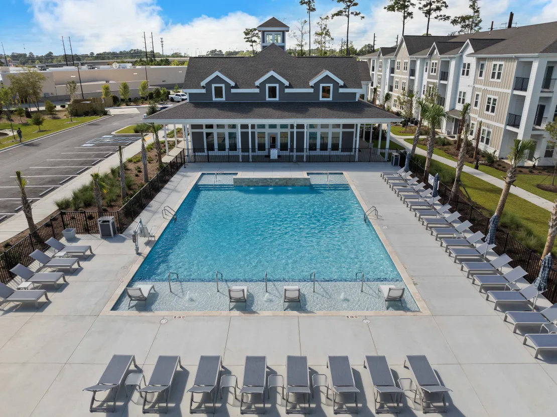 Aerial view of a large outdoor pool with lounge chairs at a modern apartment community, with a clubhouse in the background.