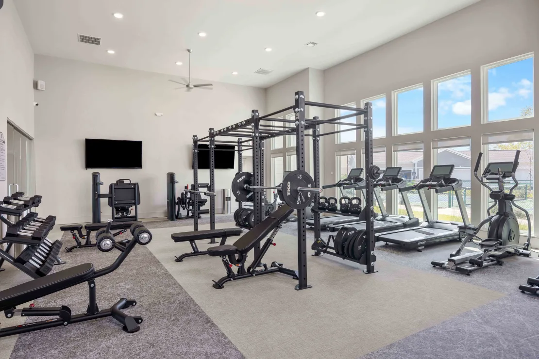 Well-equipped community gym with free weights, benches, squat rack, and cardio machines by large windows.