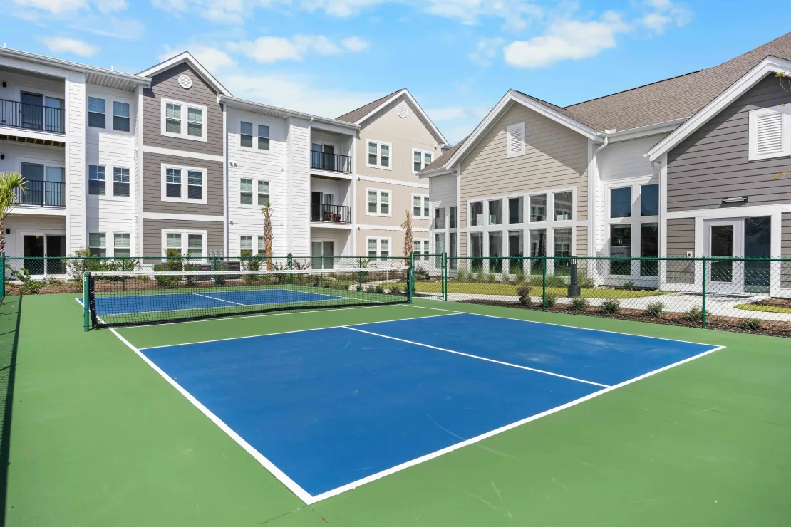 Outdoor tennis court with blue and green surface, apartment buildings in the background.