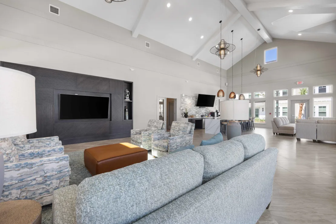 Bright, open resident lounge with blue-gray sofas, armchairs, and a wall-mounted TV.