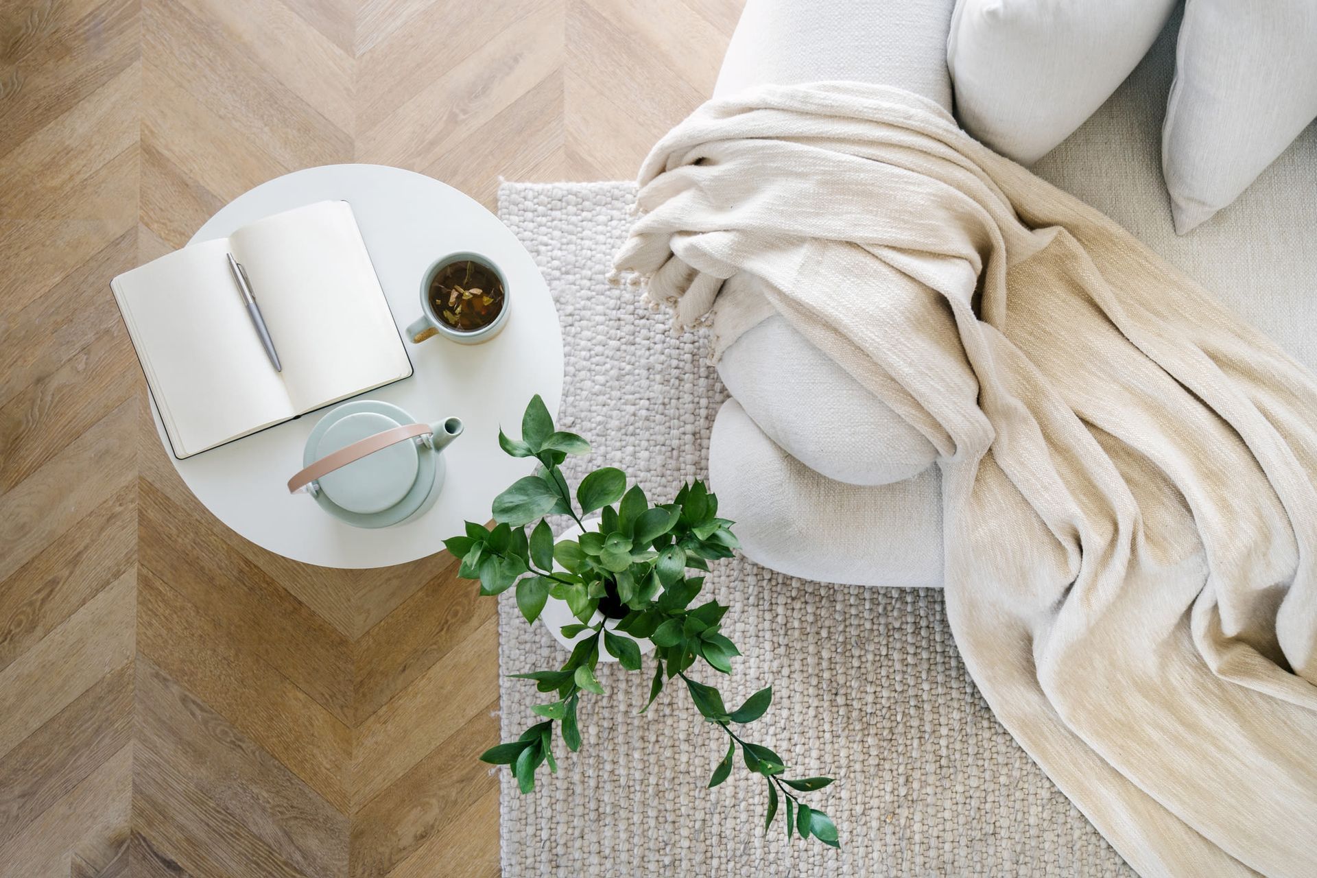 Overhead view of a cozy living room with a round coffee table, notebook, tea mug, plant, and beige blanket.