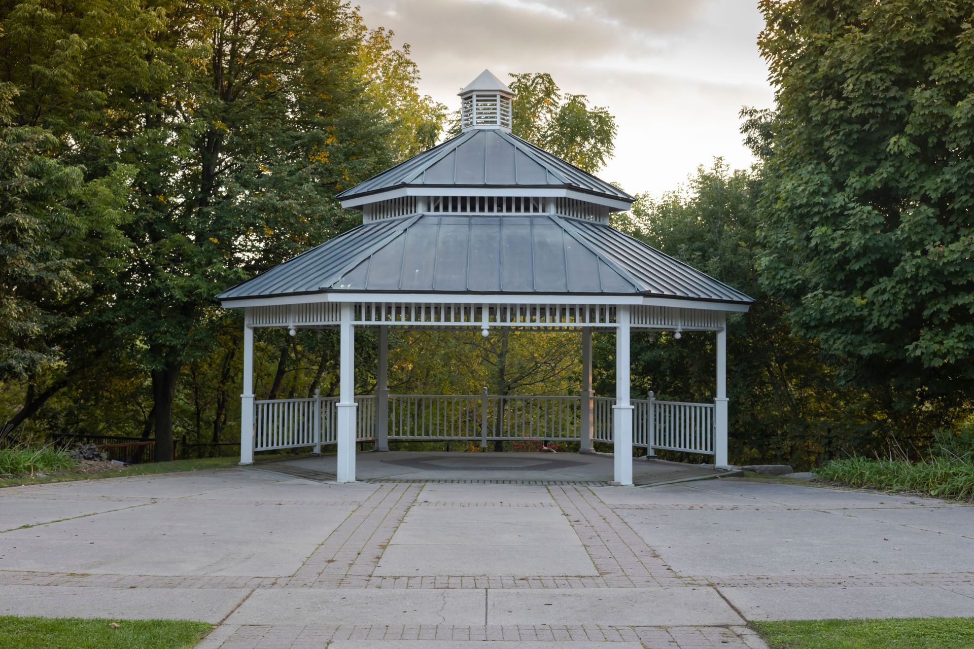 White gazebo with a tiered metal roof in a wooded park at dusk