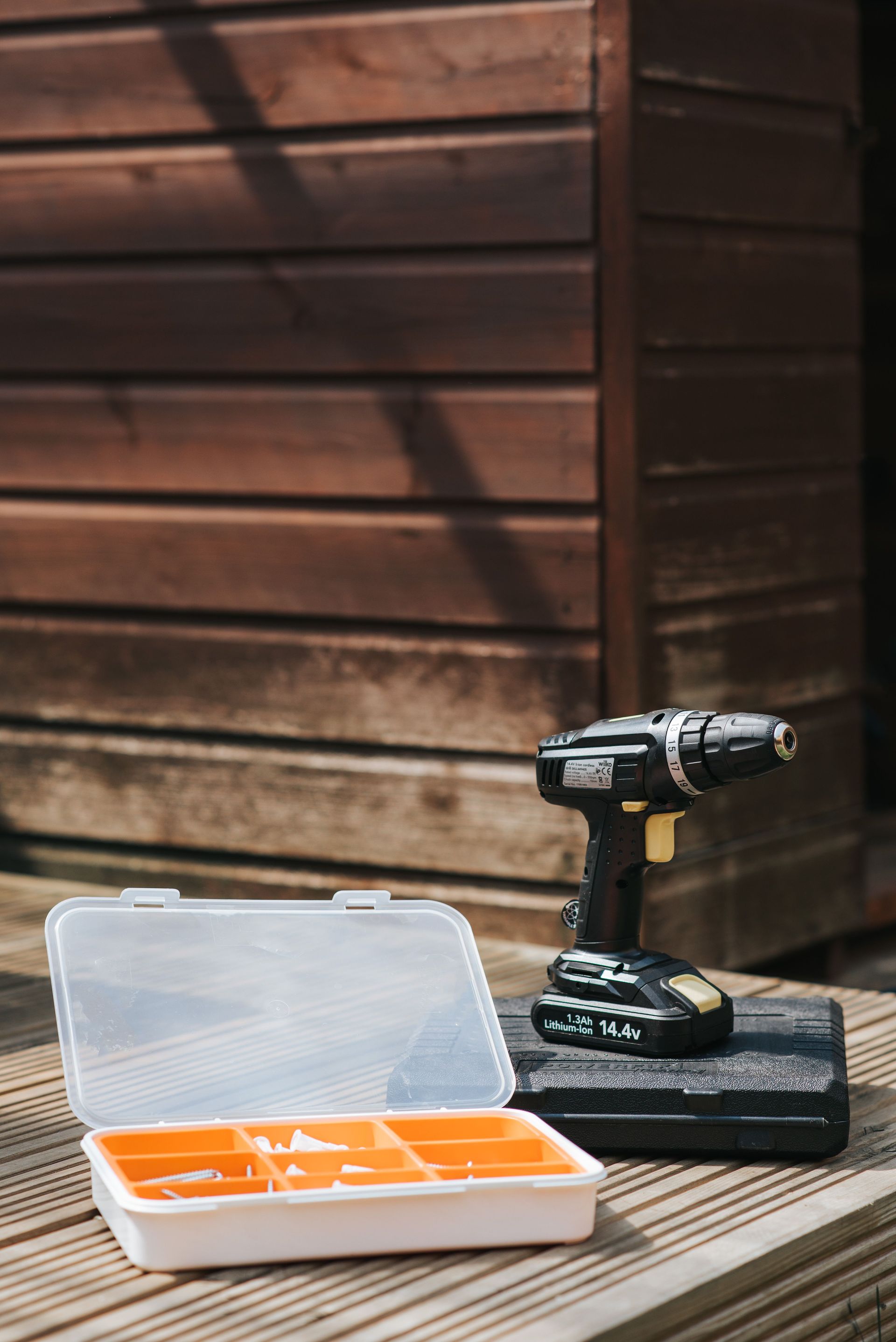 Cordless drill and drill bit case on a wooden deck beside a brown shed