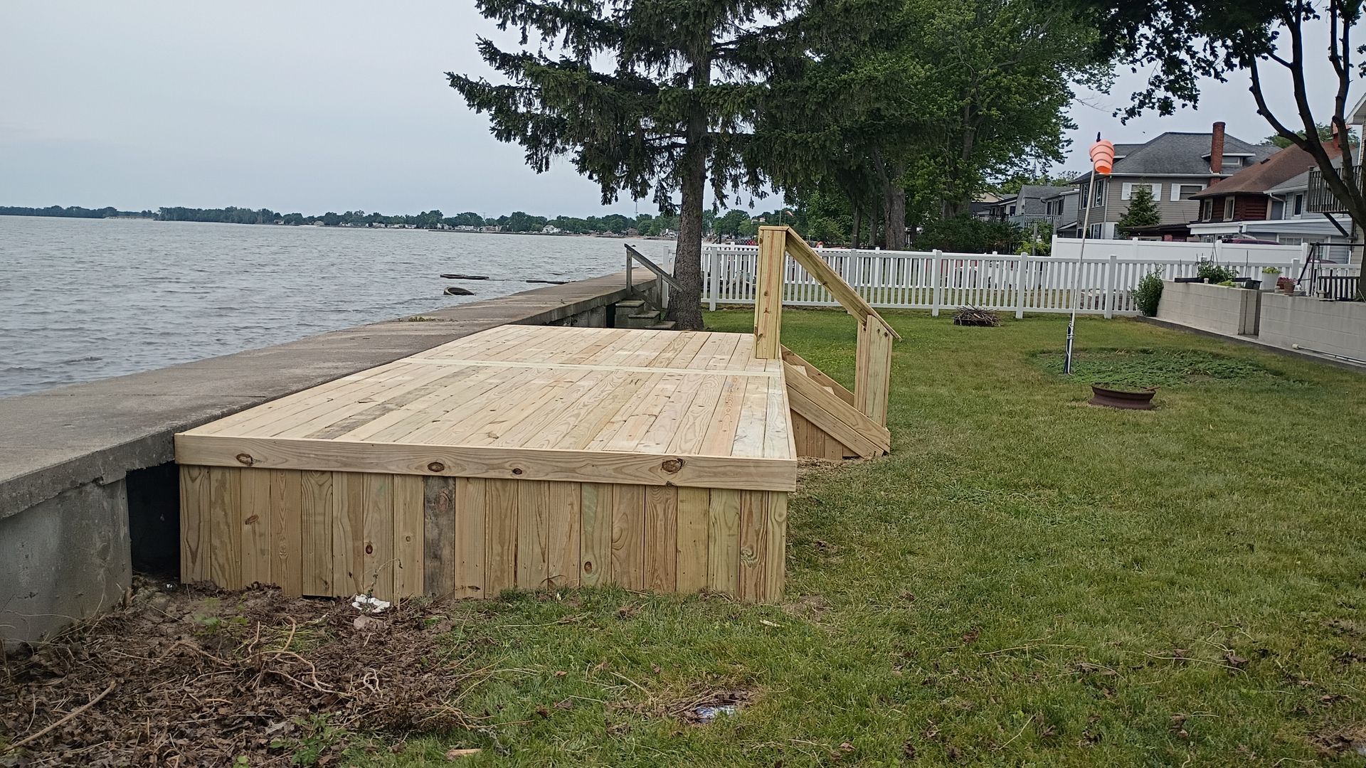 Wooden lakeside deck under construction beside grass, with a railing and water in the background.