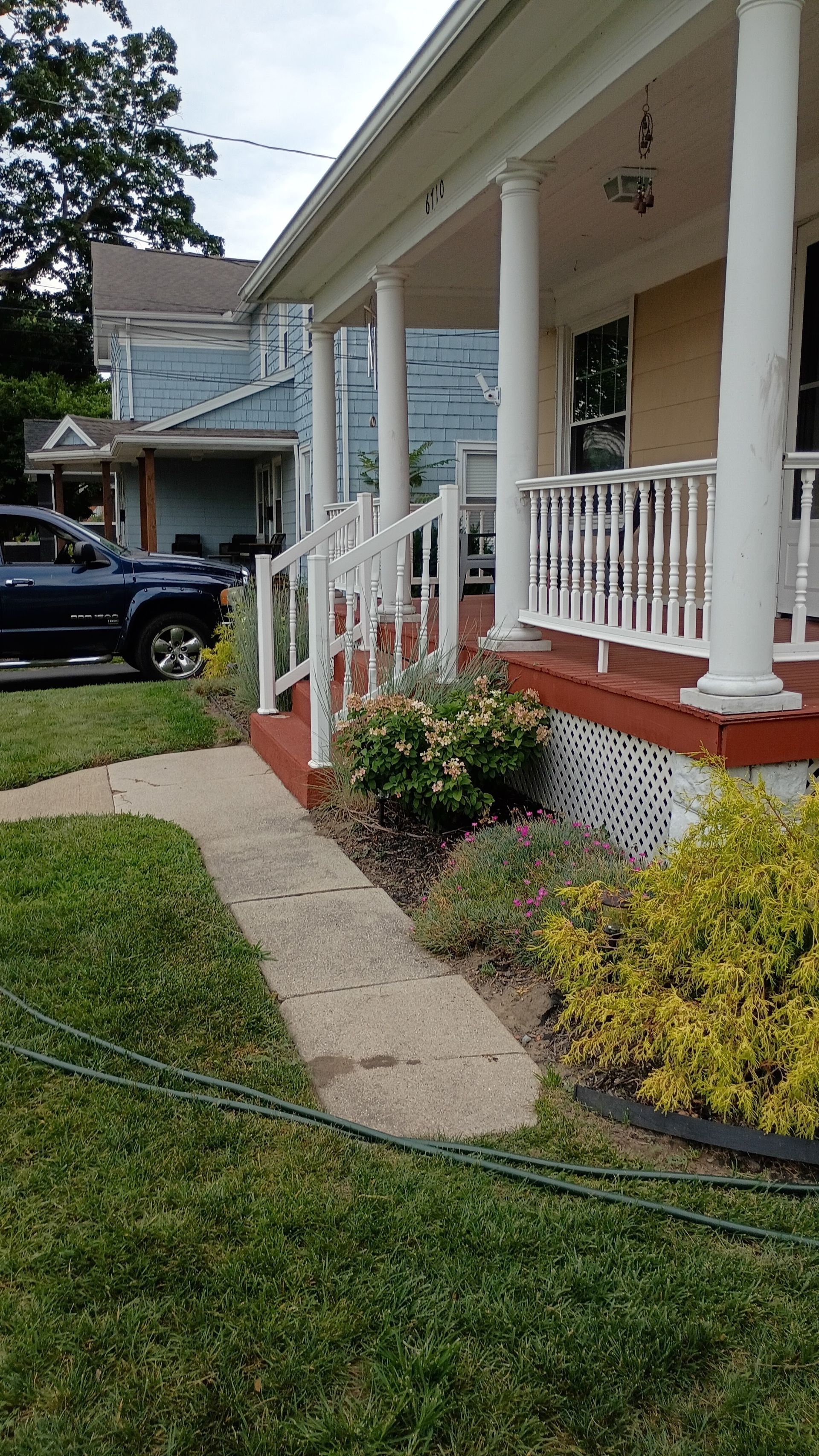 Front porch of a house with white railings, brick steps, a walkway, and shrubs in the yard.