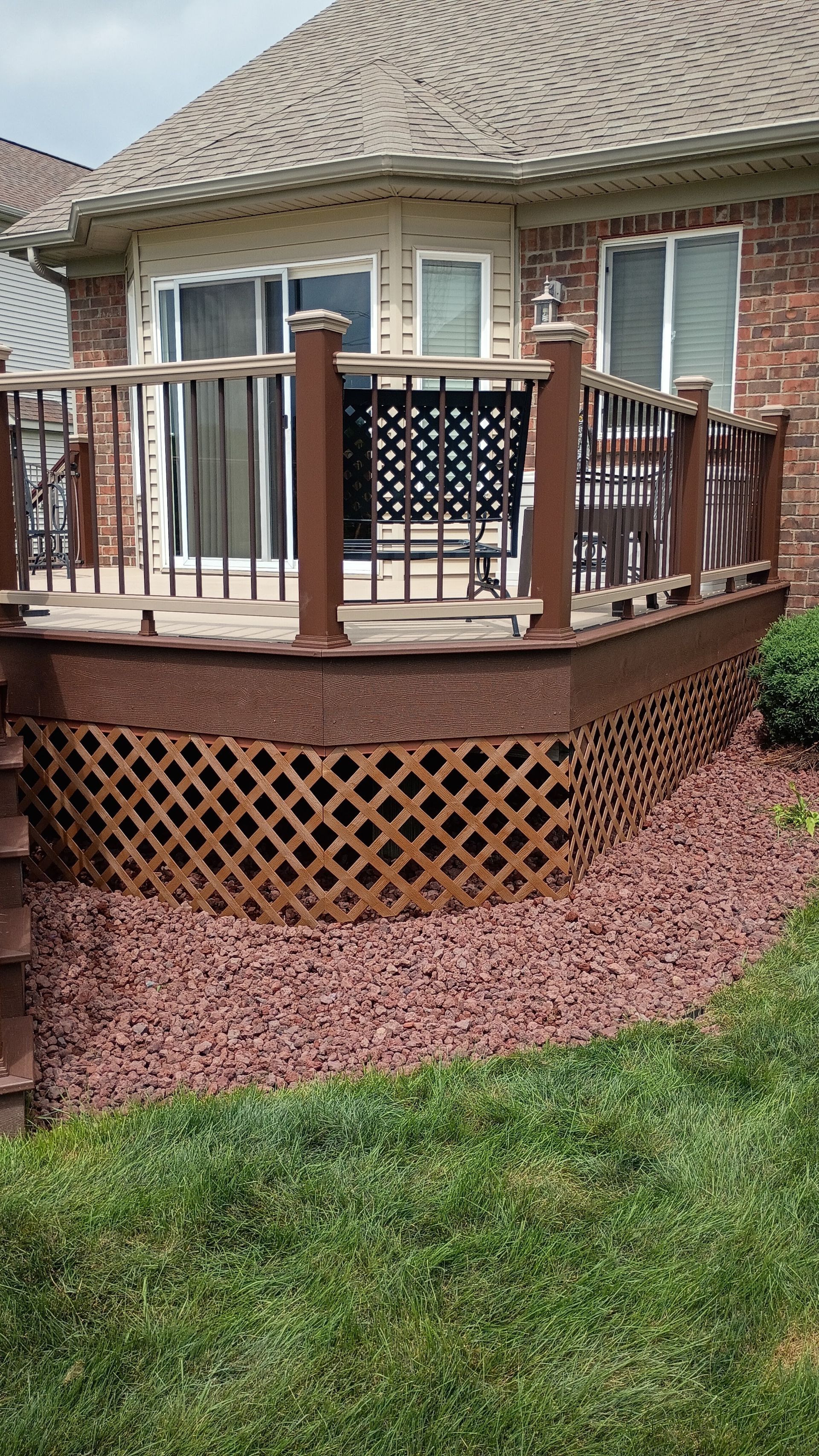 Wooden backyard deck with railing and chair beside a brick house, surrounded by red mulch and grass