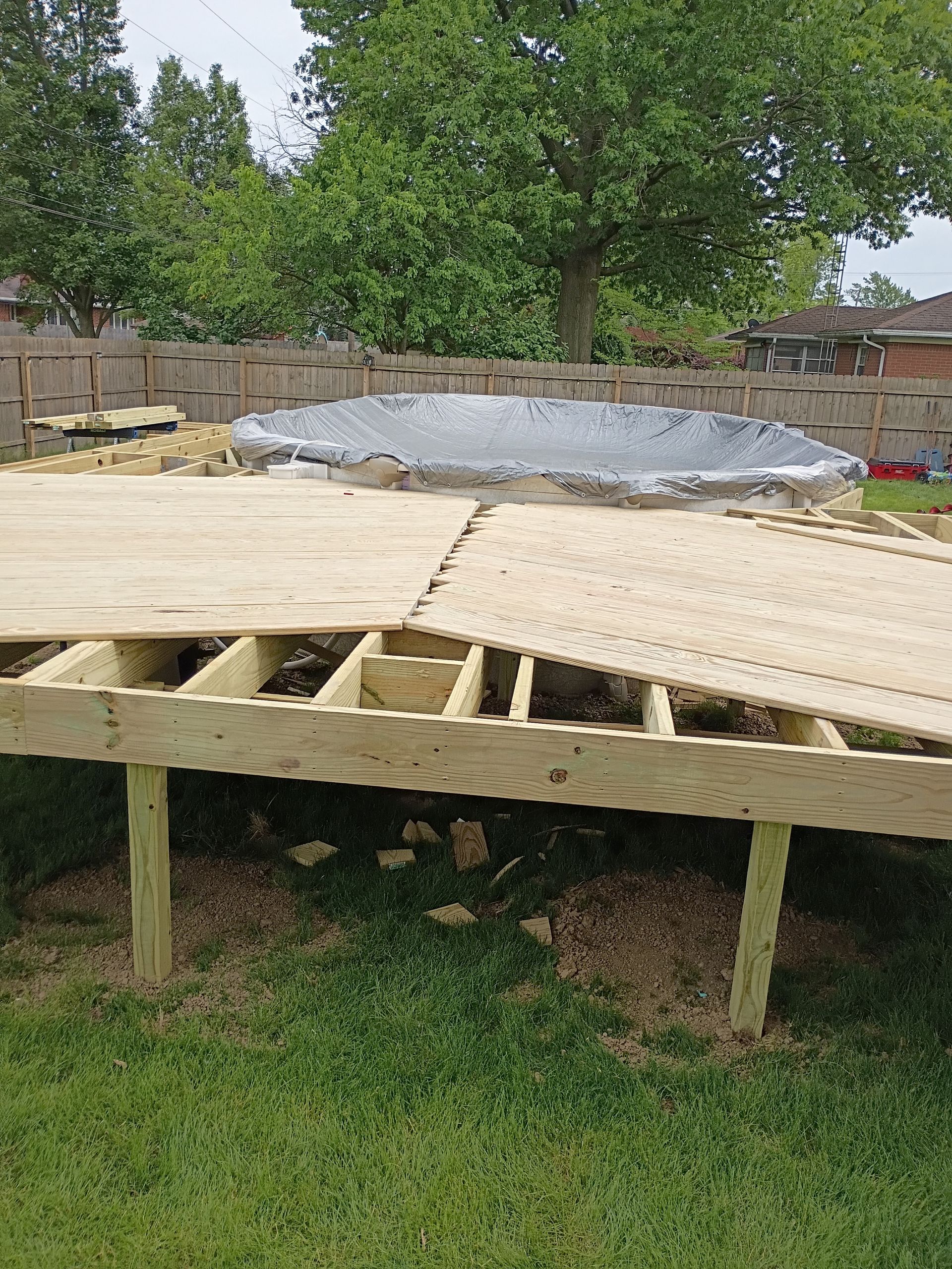 Raised wooden deck under construction in a backyard, with a tarp-covered mound and trees behind it