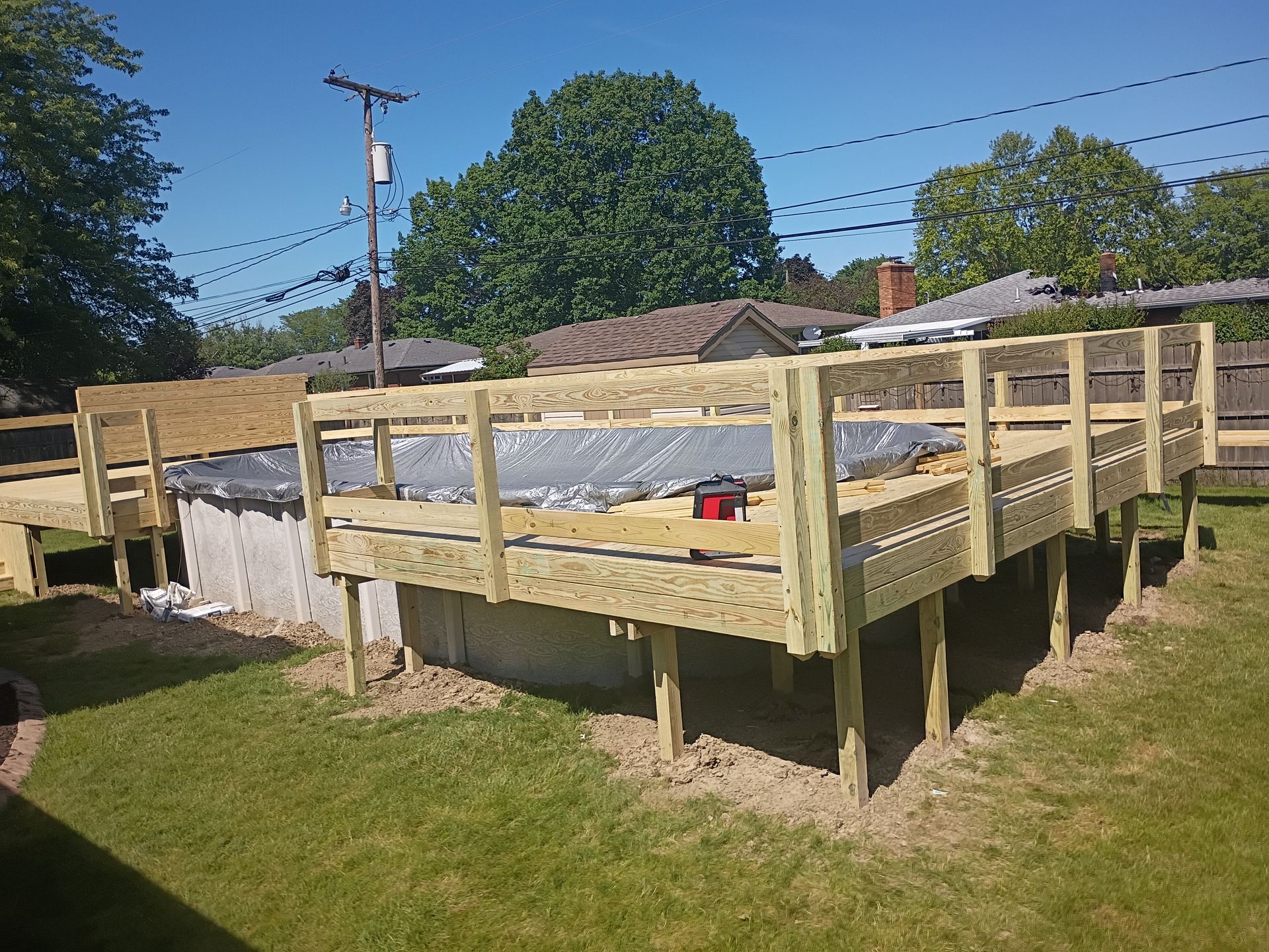 Wooden backyard deck under construction on posts, surrounded by grass and trees, with a house in the background