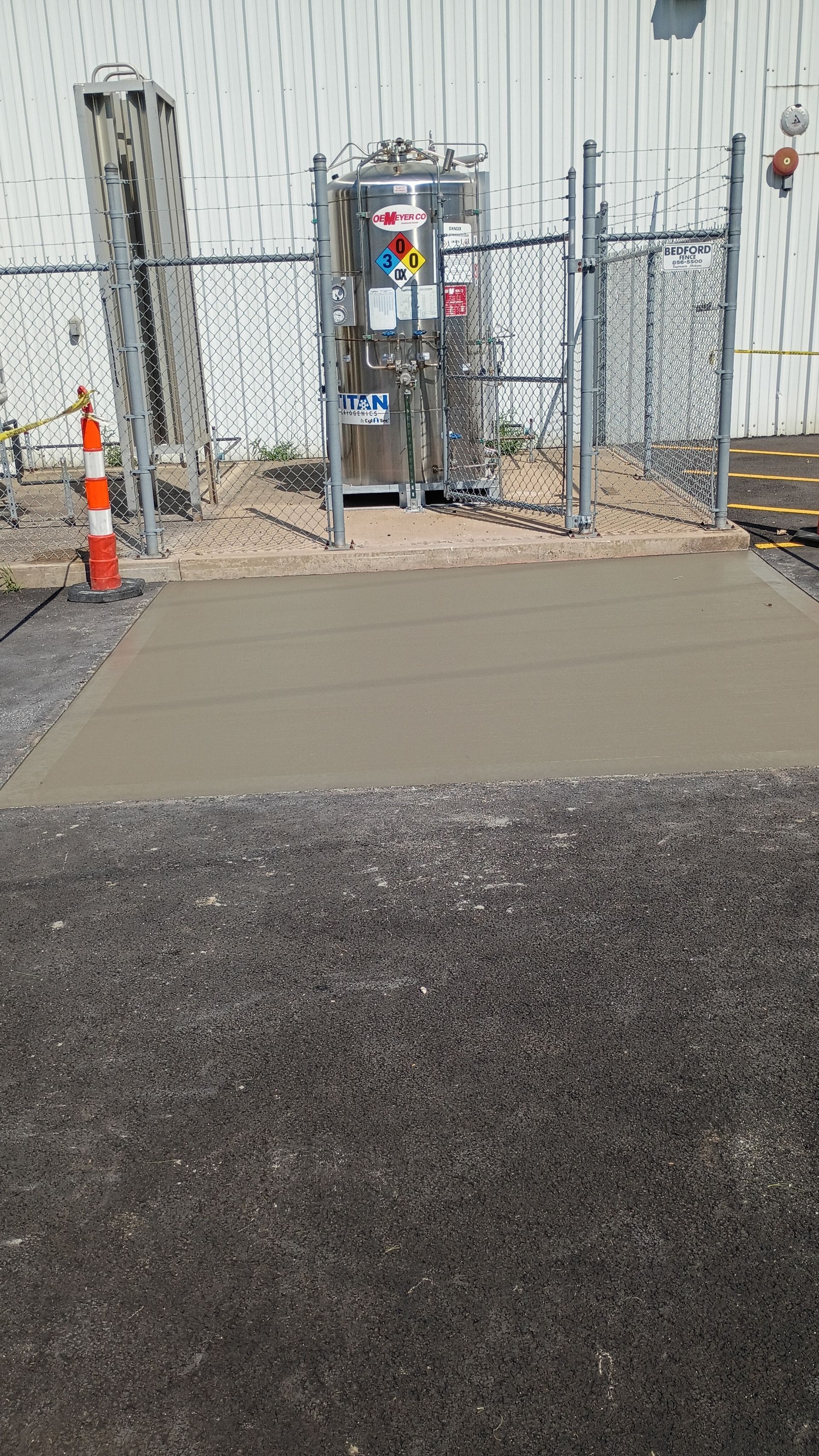 Concrete walkway and fenced utility area beside a building, with traffic cones and equipment.