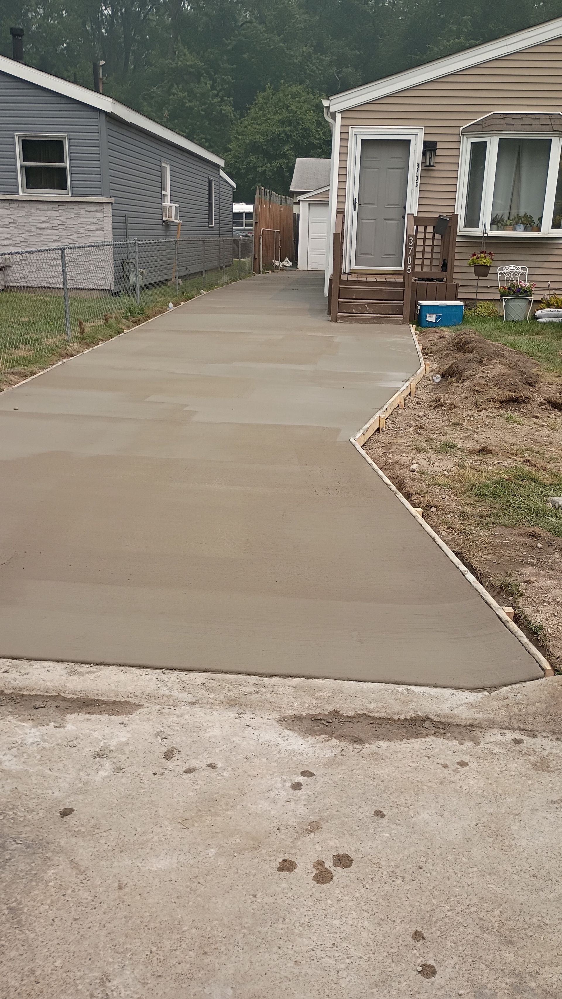 Freshly poured concrete walkway between two houses, leading to a driveway and front porch area.