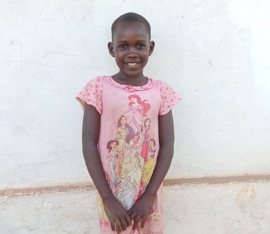 Girl in a pink dress with a cartoon print smiles in front of a white wall.