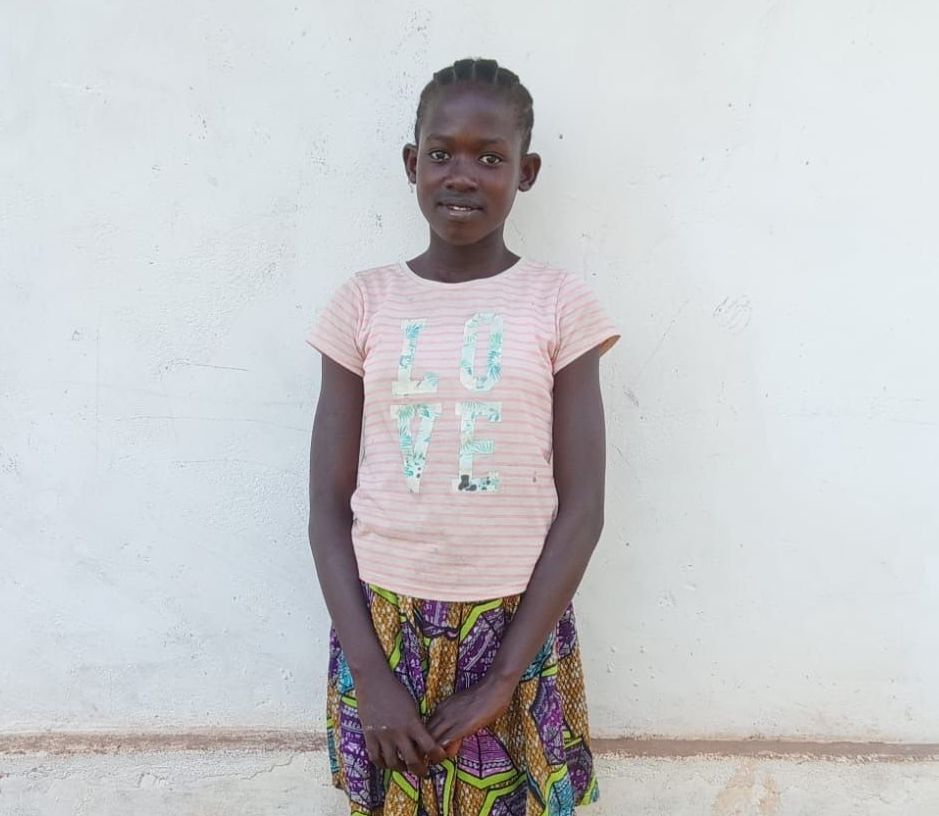 Girl standing in front of a white wall, wearing a pink shirt and patterned skirt, smiling.