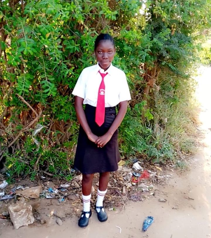 Girl in school uniform stands outside by bushes.