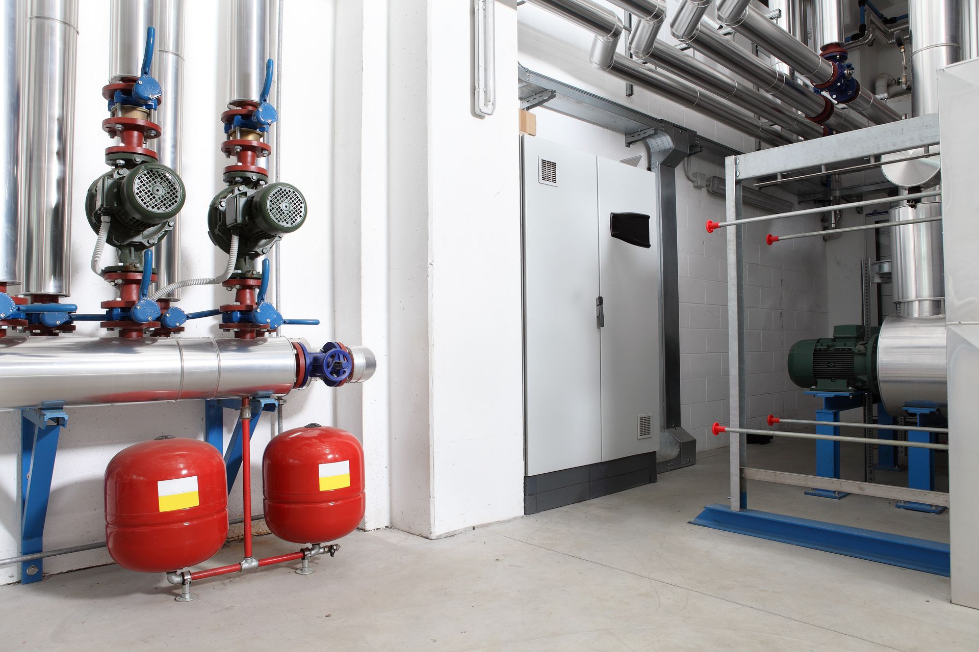 Industrial boiler room with pipes, valves, red expansion tanks, and a large pump system.