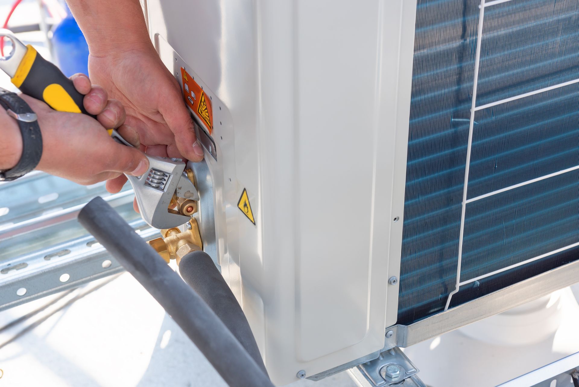 Hands using tools to service an outdoor air conditioning unit, with hoses and exposed components visible.