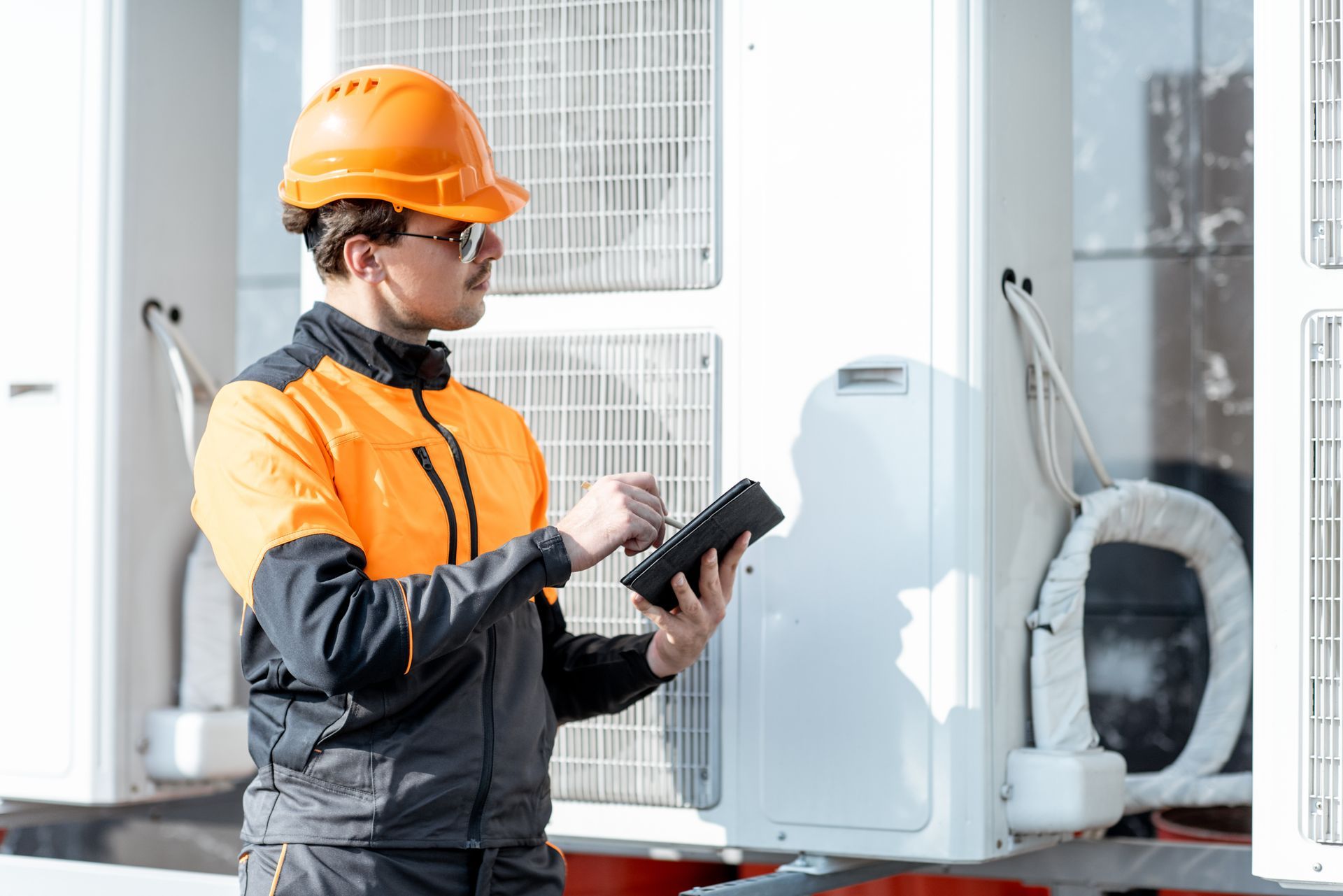 Technician in orange hard hat using a tablet beside industrial equipment outdoors