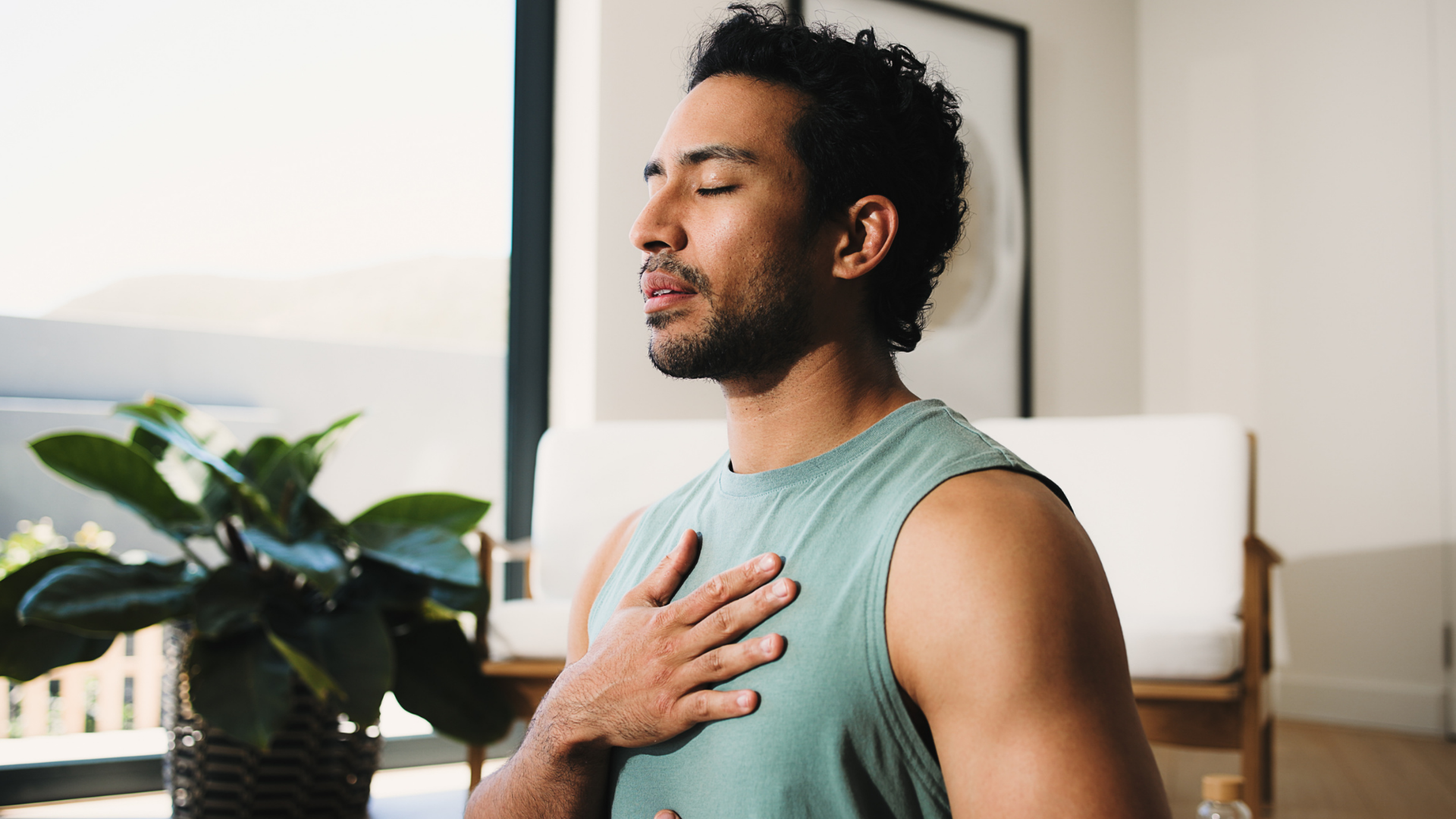 Man in a teal sleeveless shirt sits indoors with eyes closed, hand on chest, taking a deep breath.