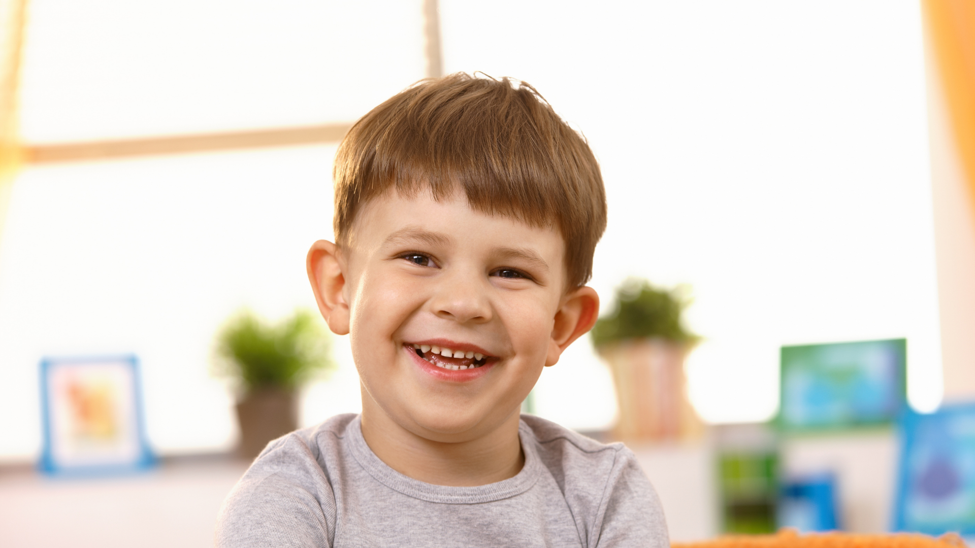 Smiling child in a gray shirt in a bright classroom with colorful blurred shelves in the background