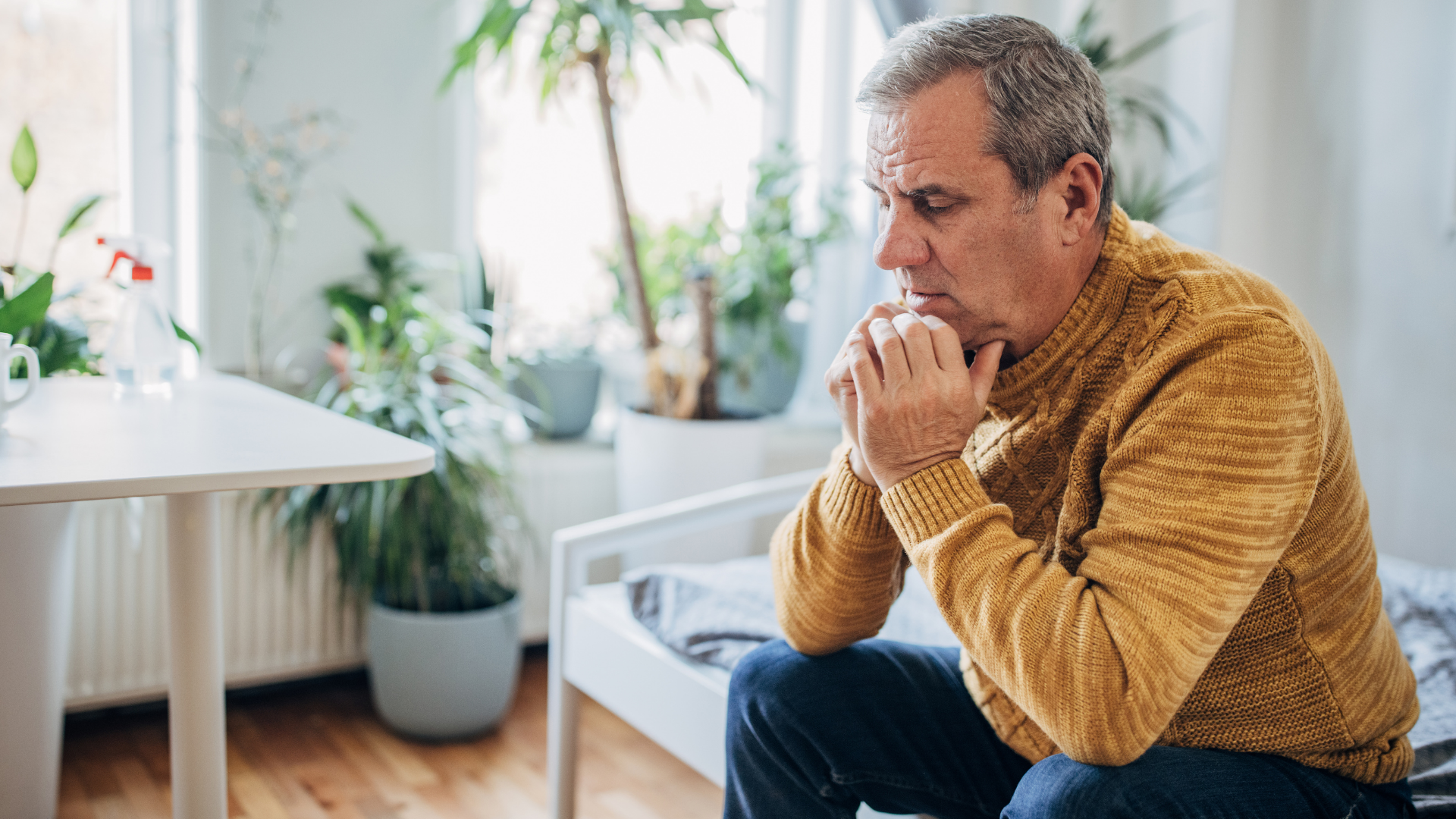Man sitting on a couch with hands clasped, looking thoughtful in a bright living room with plants.