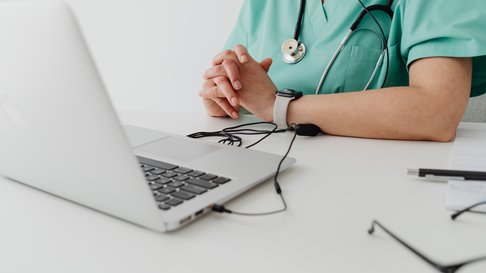 Doctor in green scrubs with stethoscope typing beside a laptop at a desk in a bright office