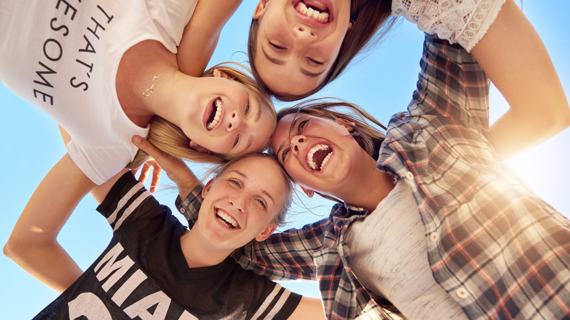 Four friends smiling in a circle against a blue sky, arms around each other, viewed from below.