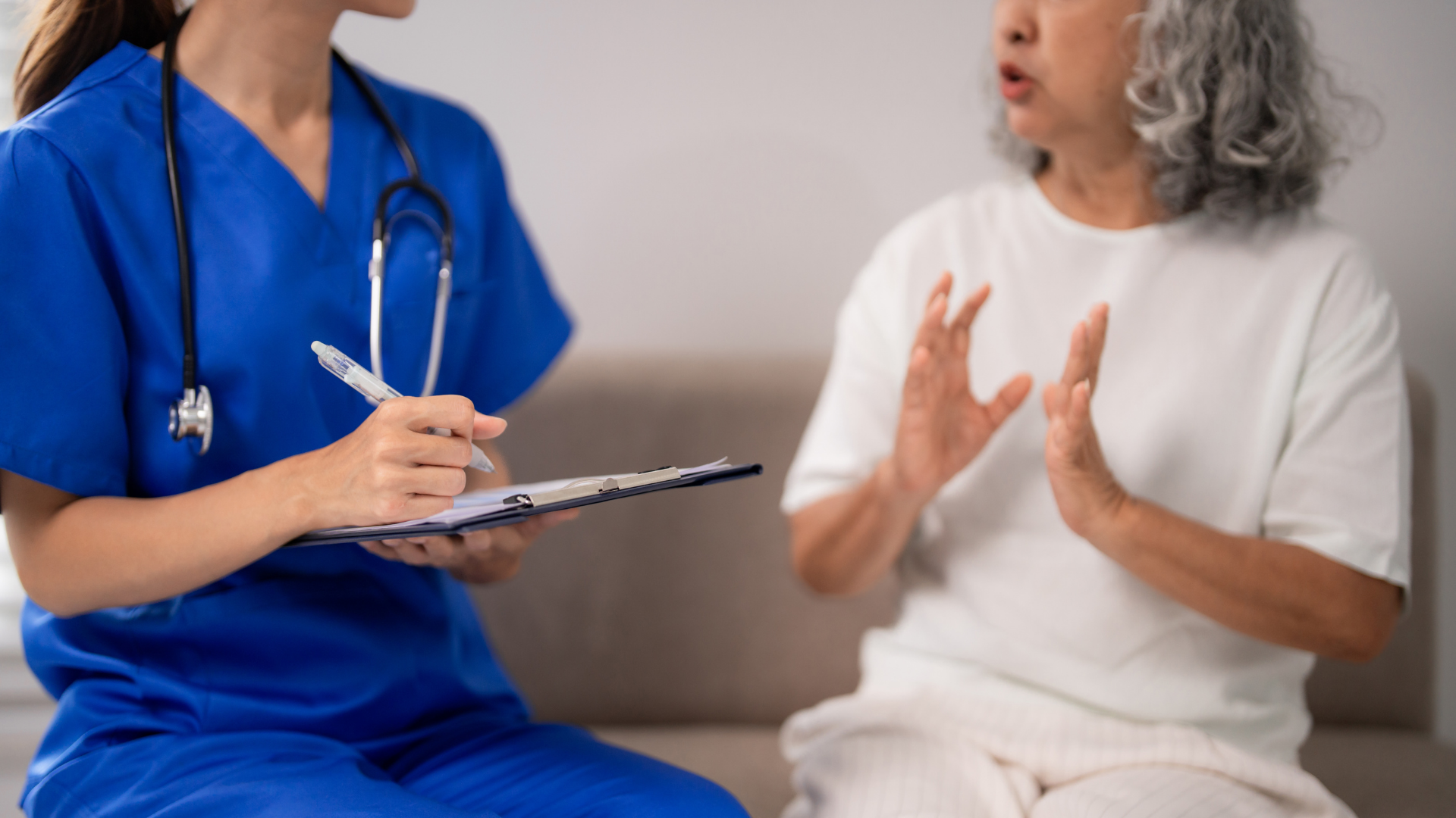 Nurse in blue scrubs taking notes while speaking with a patient on a couch in a home setting