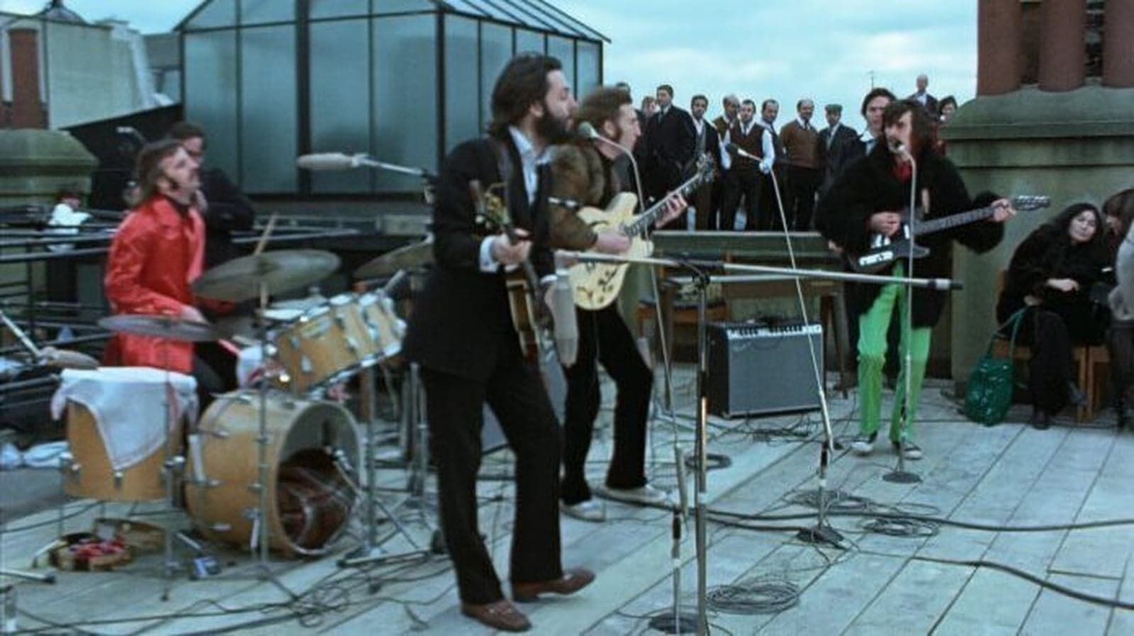 Band performing on a rooftop deck with drums, guitars, and microphone stands, audience gathered nearby.