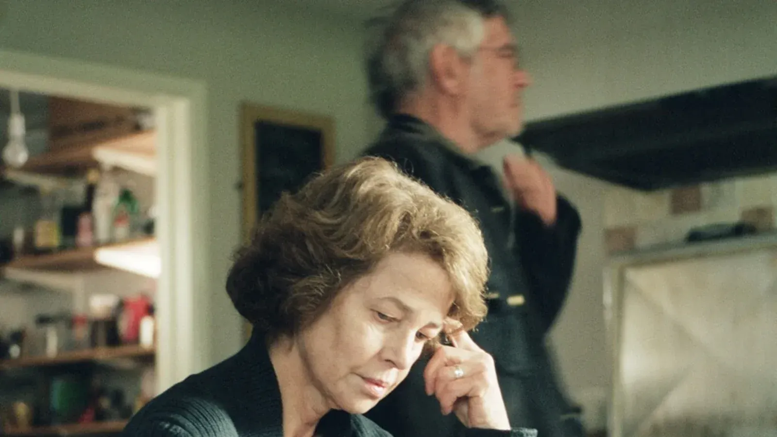 Woman with short hair, hand to forehead, in kitchen; man in background.