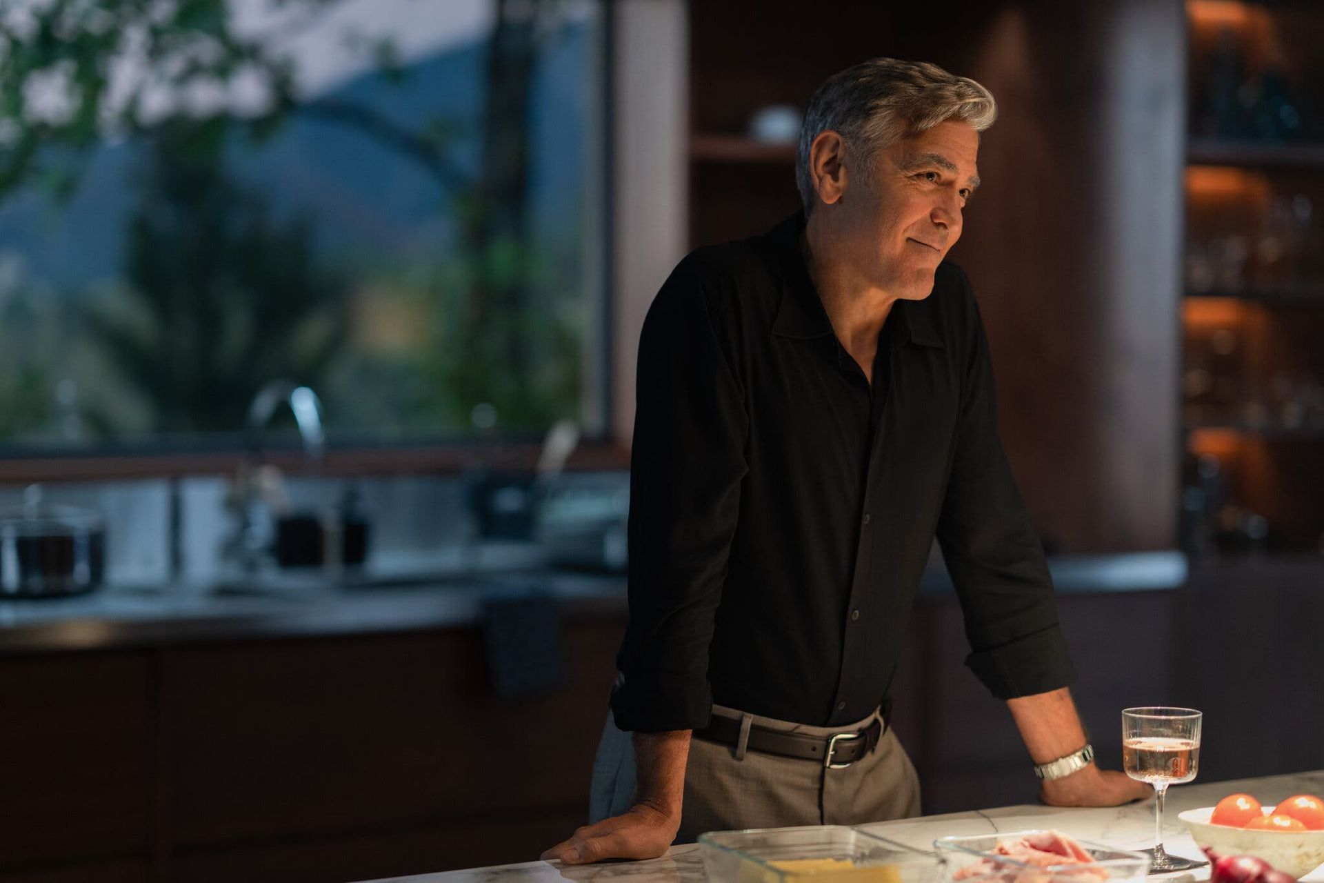 Man leaning on a kitchen island beside plated food, with a drink in a modern, softly lit kitchen.