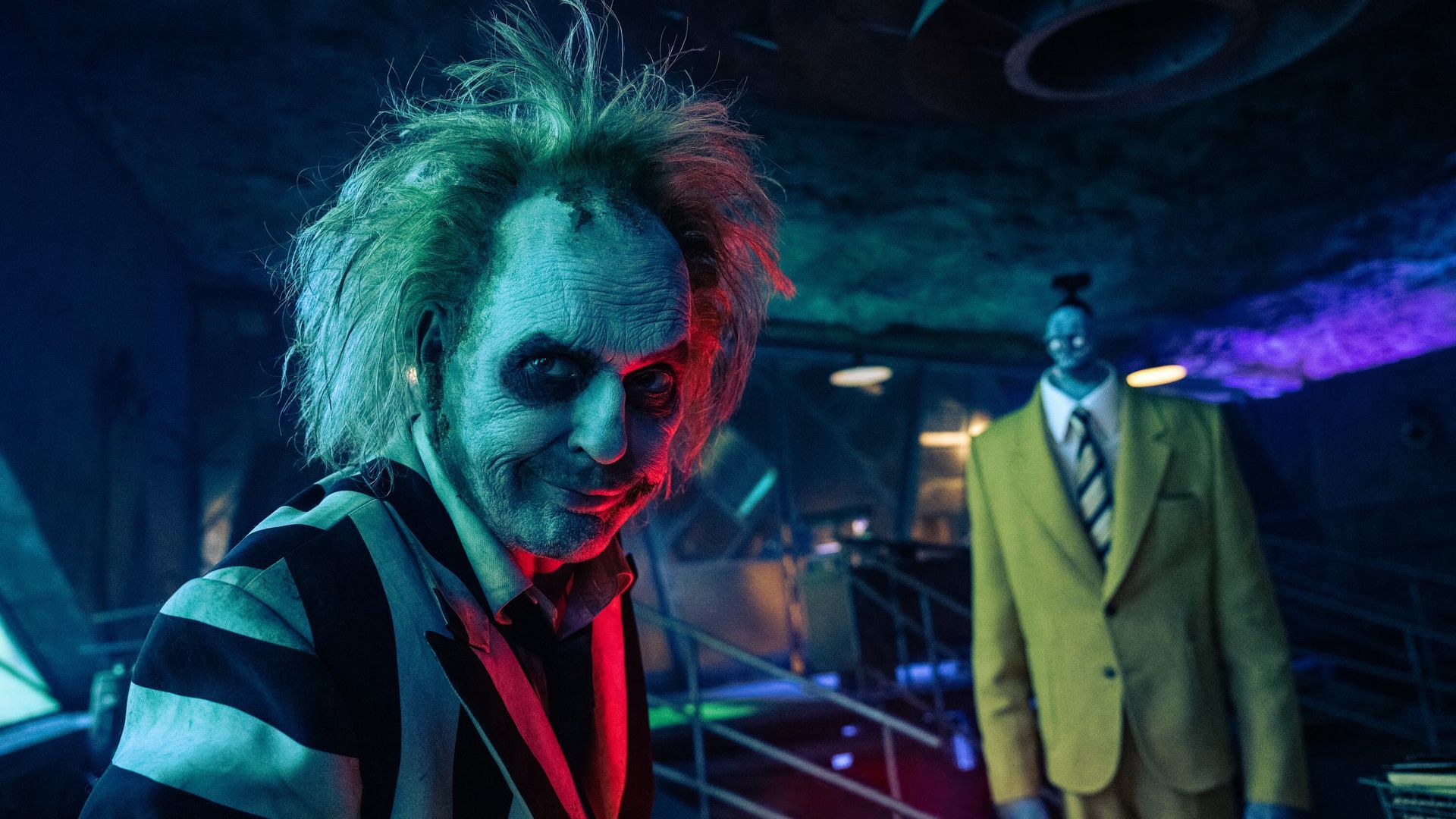 Pale-faced figure in green hair and striped suit in a neon-lit room, with a yellow-suited man behind.