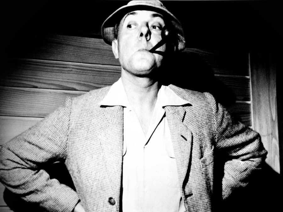 Black-and-white photo of a person in a hat, hands on hips, smoking a cigarette indoors