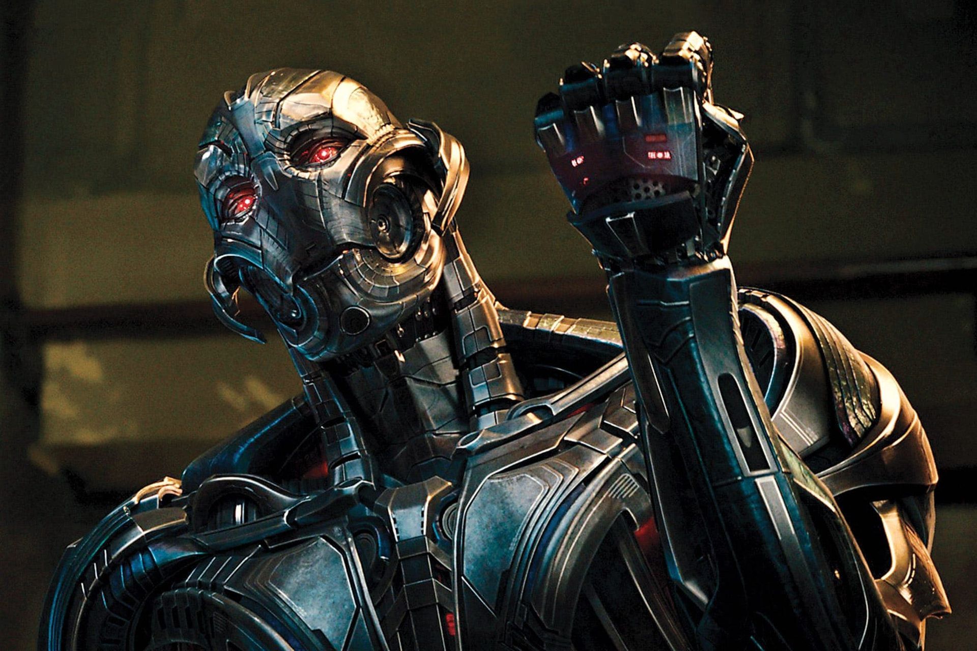 Silver robot with glowing red eye raises a clenched fist in a dark industrial setting