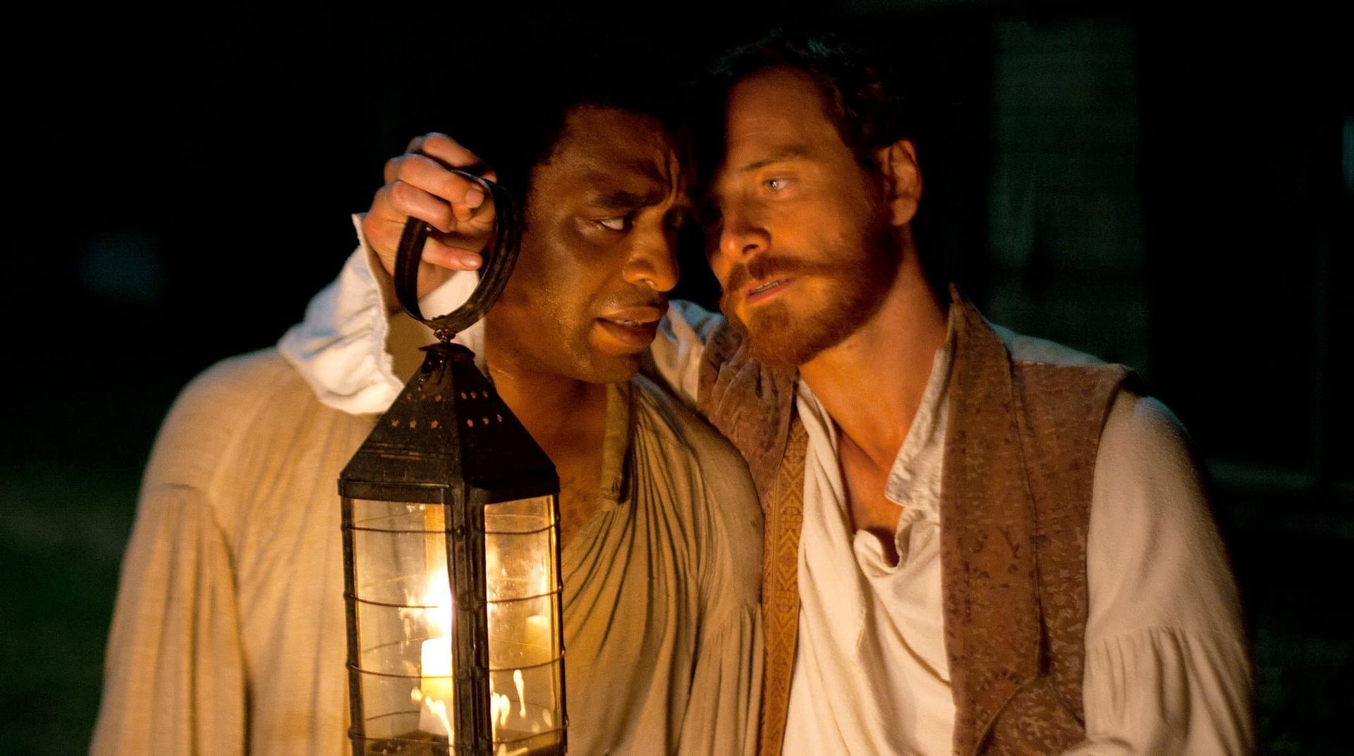 Two men in period clothing share a lantern in a dark scene, one leaning close and whispering.