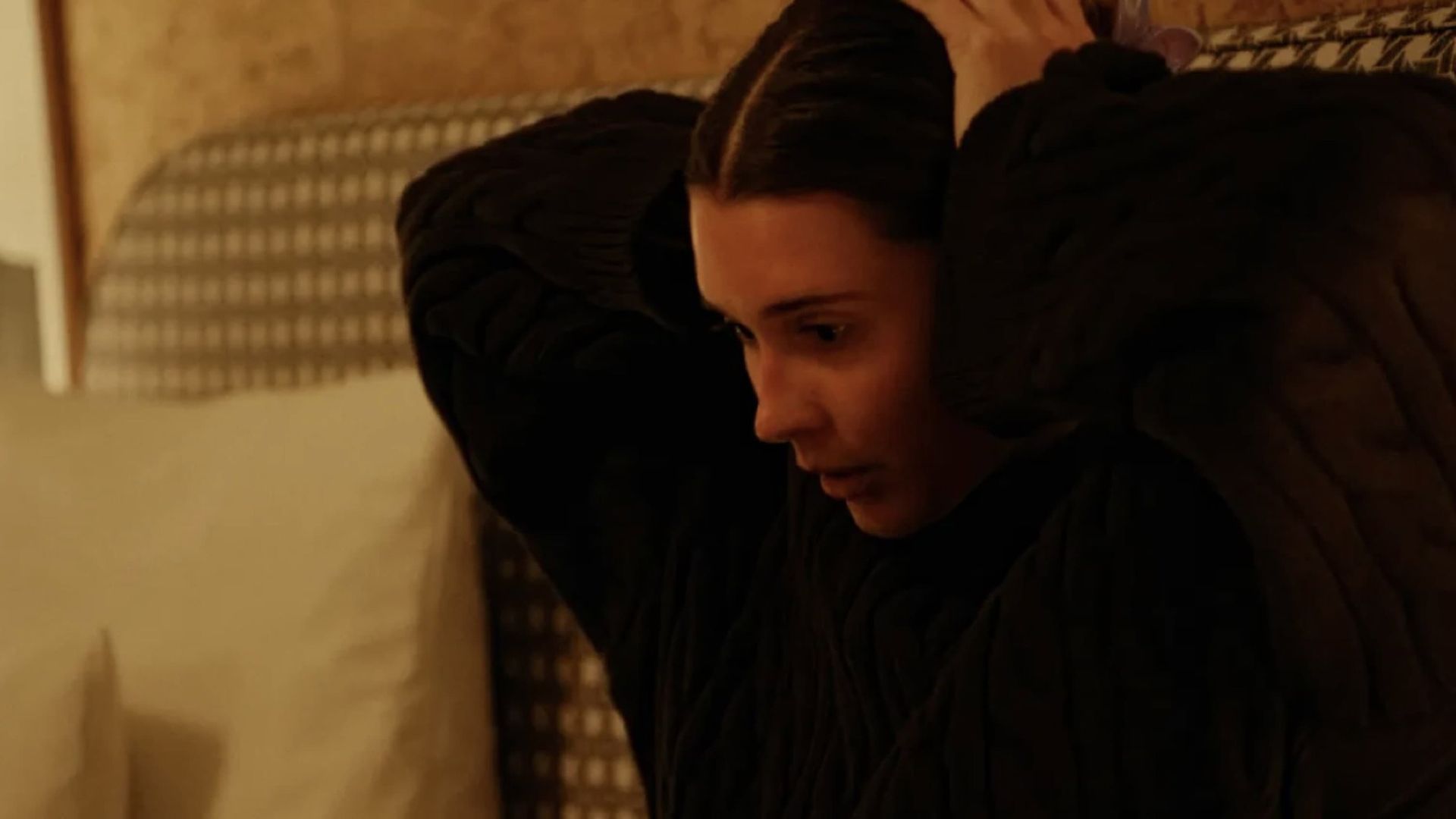 Person in dark clothing with hands behind head, sitting on a bed in a warm-lit bedroom.