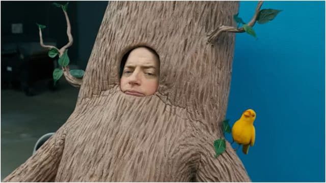 Woman peeking from a tree costume beside a small yellow bird on a blue backdrop