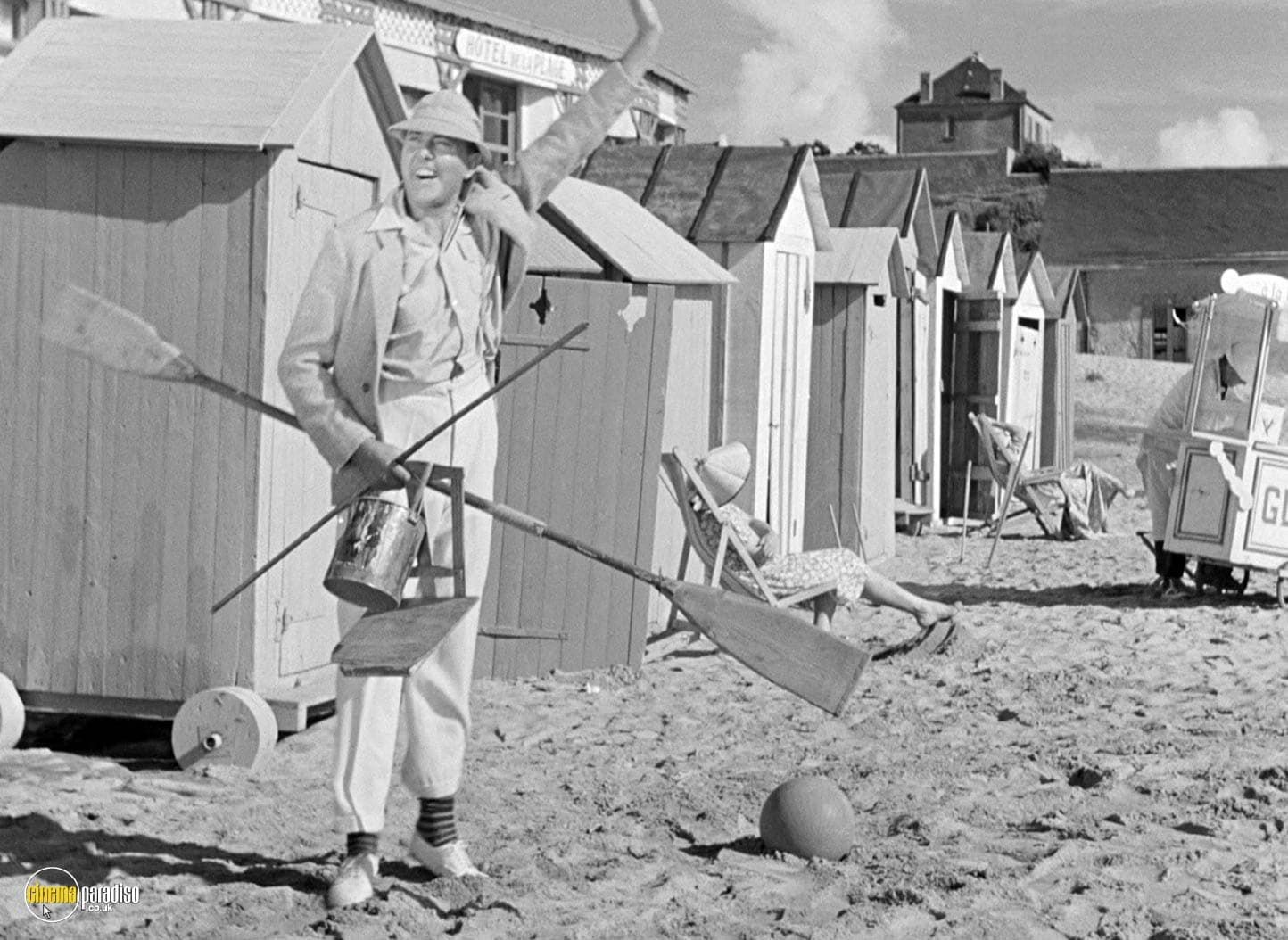 Man holding garden tools in front of beach huts on a sandy shoreline