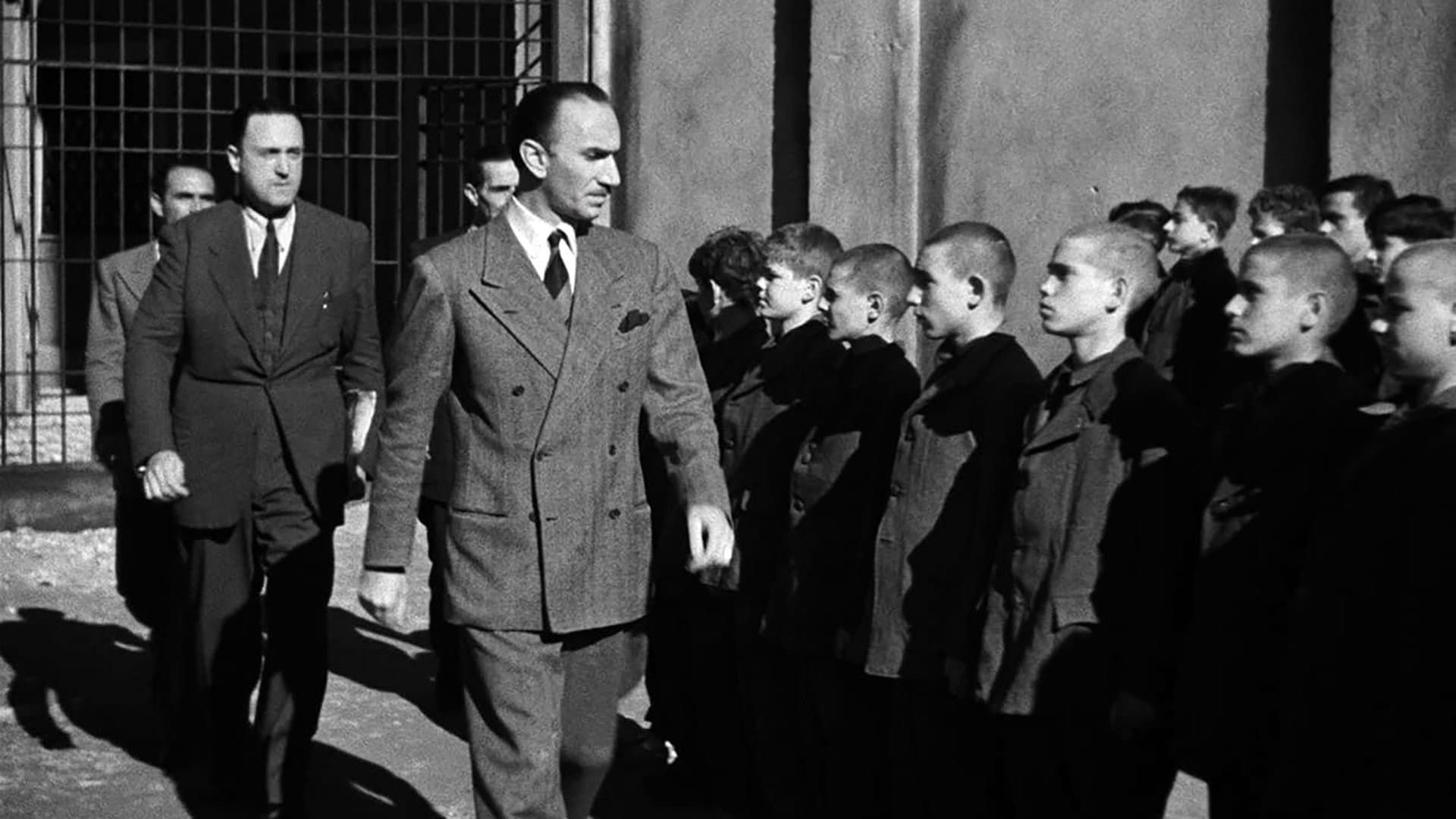 Officials in dark suits inspect a line of children standing at attention outdoors by a barred gate.