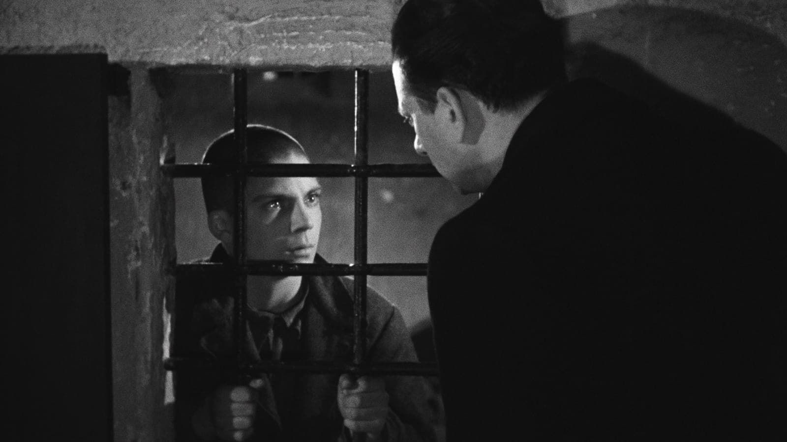 Two people talk through barred windows in a dim black-and-white room