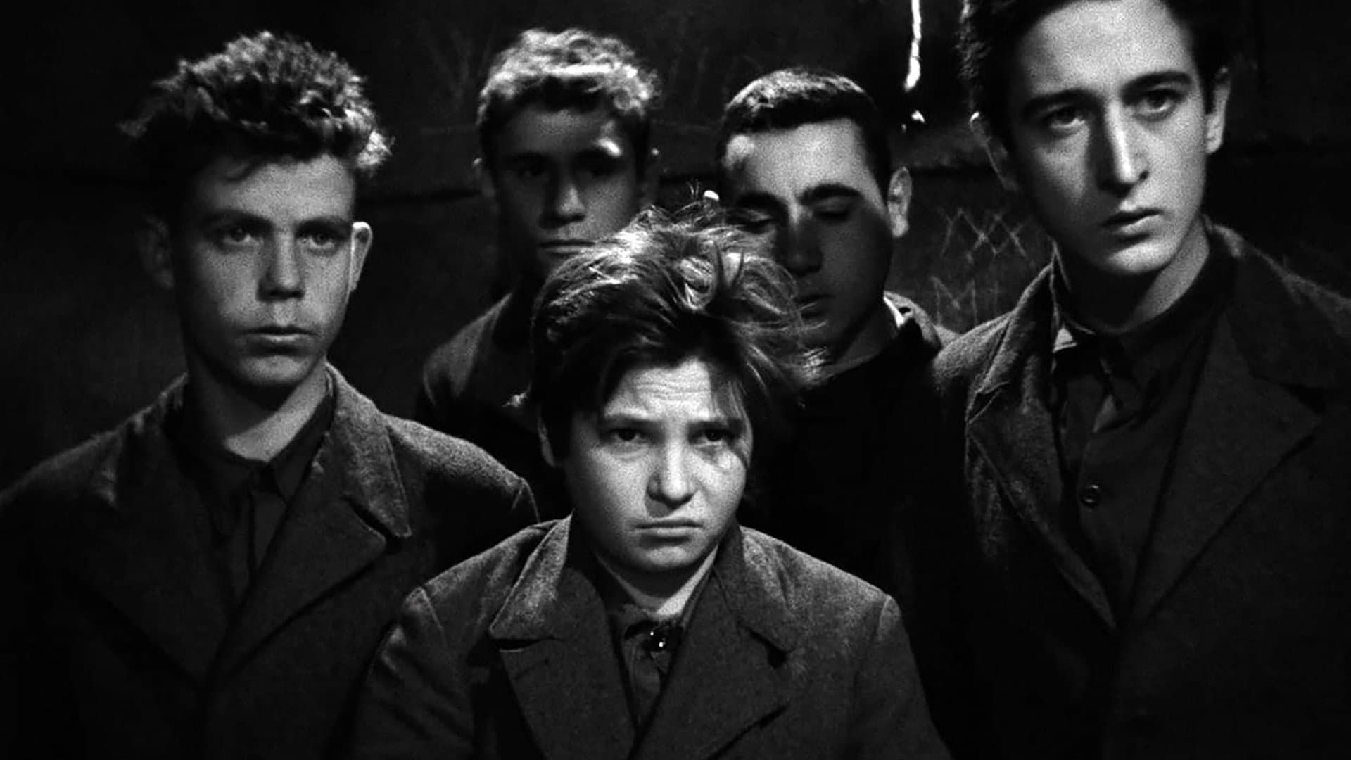 Five people in dark clothing pose in dramatic black-and-white lighting, facing the camera.