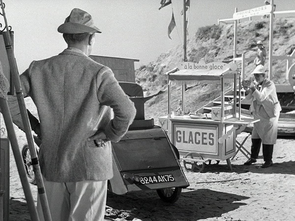 Street scene with a man by a car, an ice cream cart, and a vendor serving in the background.