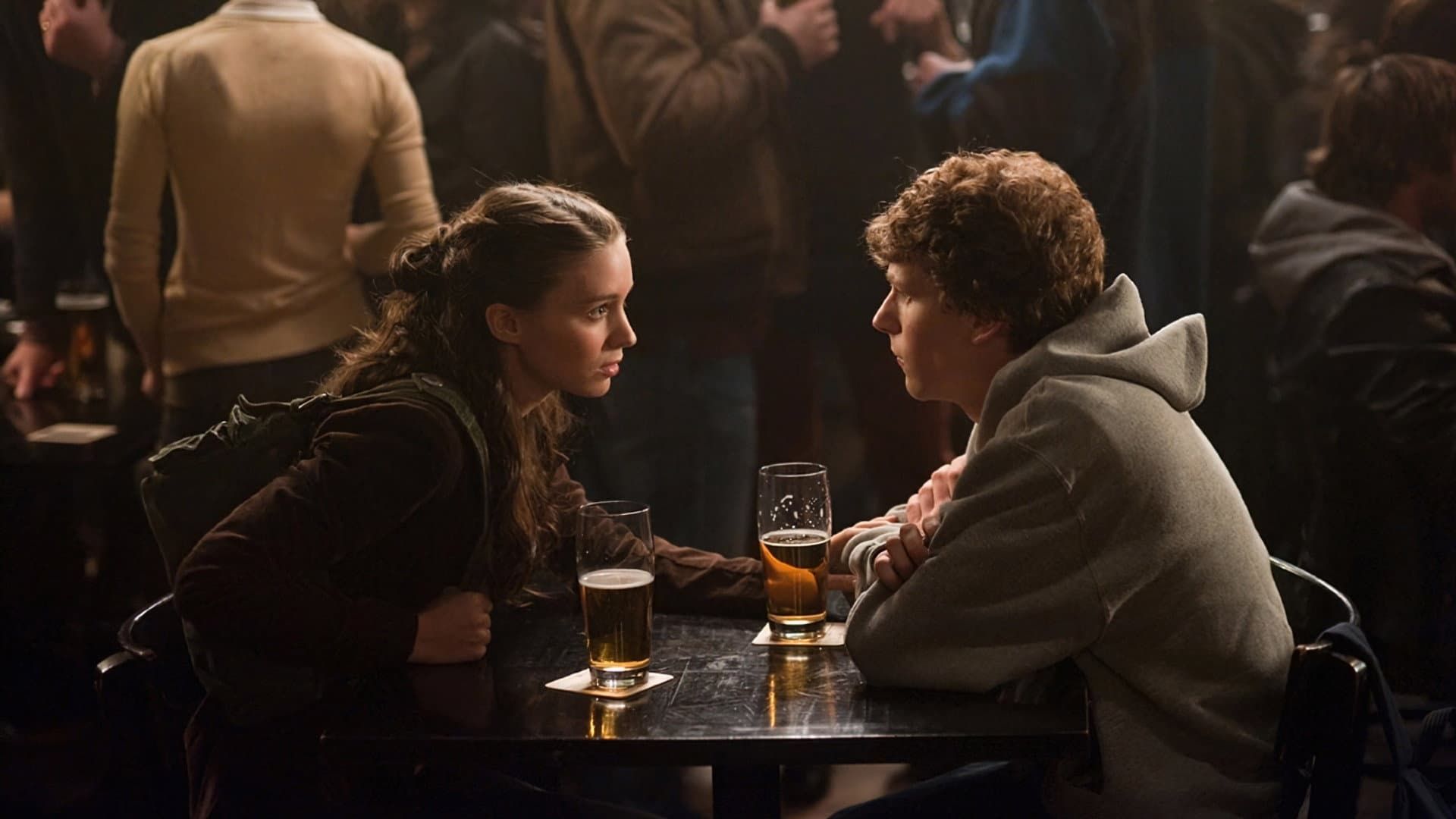 Two people leaning across a pub table, talking over drinks in a dimly lit bar.