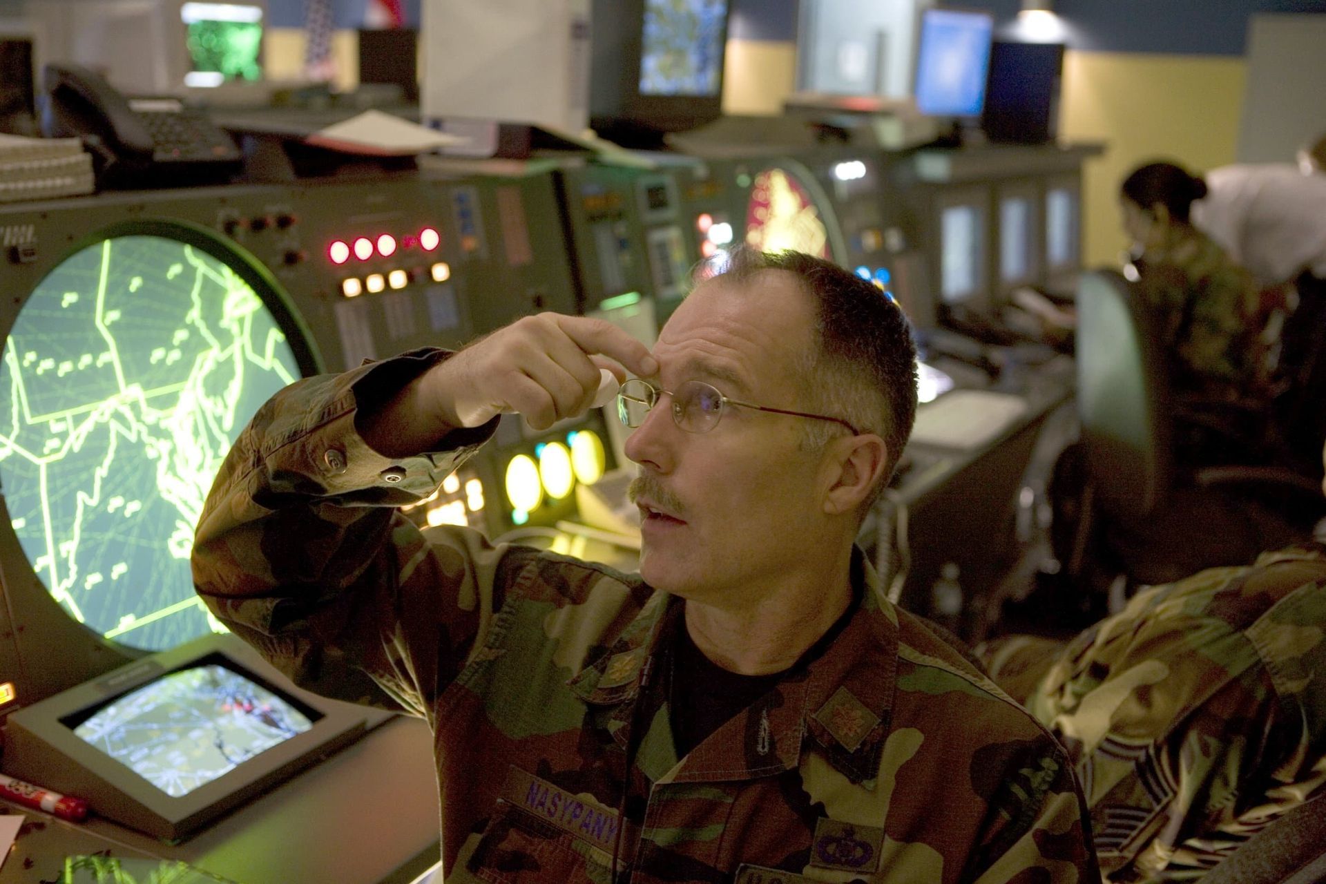 Person in military uniform using a headset in a dim control room with glowing radar screens