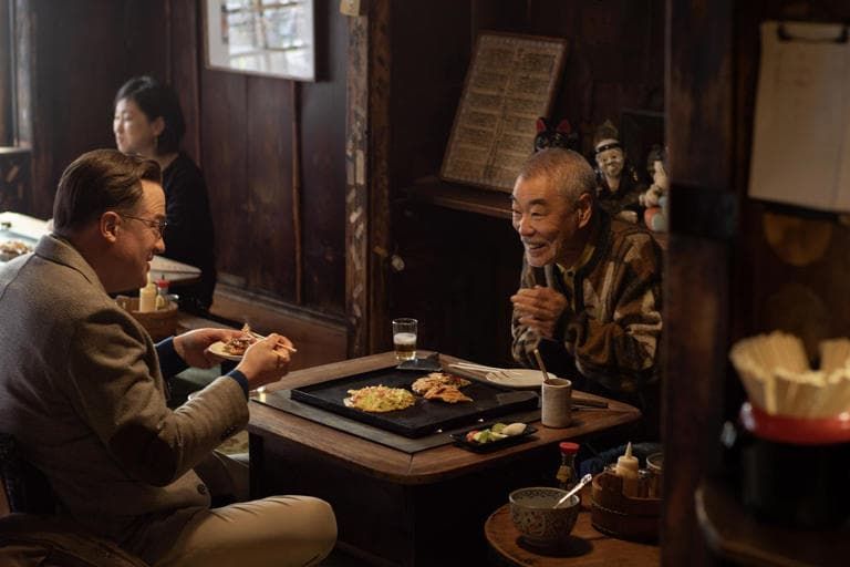 Two people dining at a dimly lit restaurant, one smiling across a small table with food and drinks.