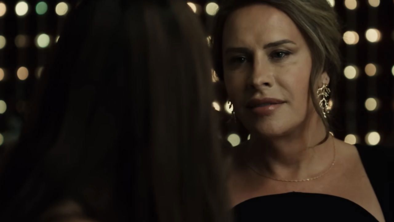Woman in a black dress talking to a person, likely at a party. Her expression is serious.