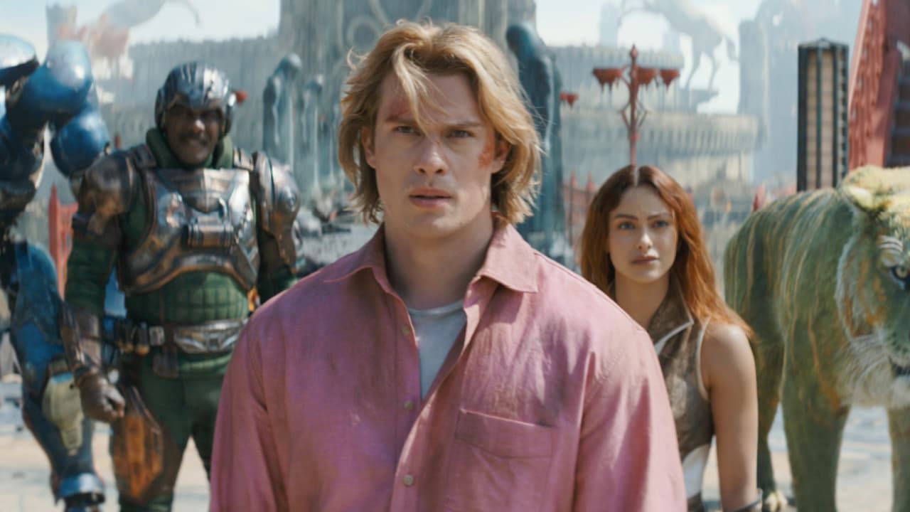 A man in a pink shirt stands in front of a woman and a robot in a dusty outdoor setting.