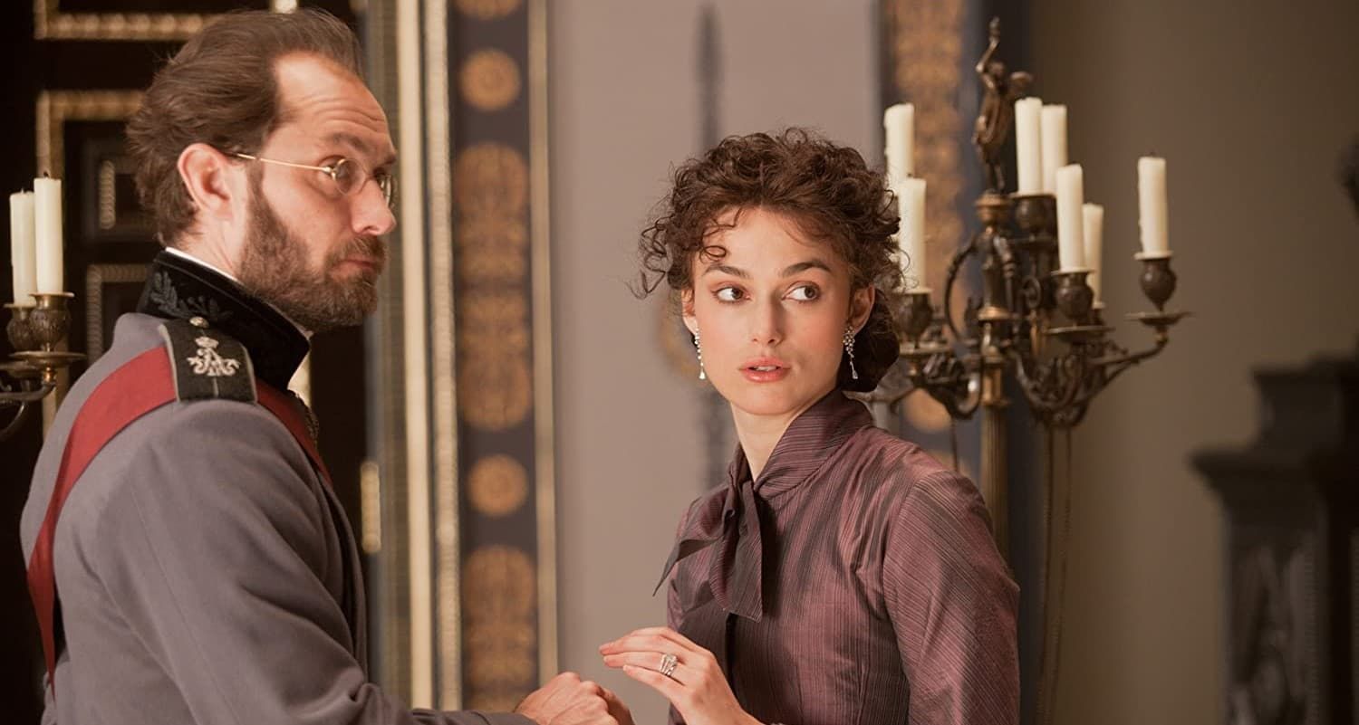 Two people in period clothing stand indoors near a candelabra, one looking back with concern.